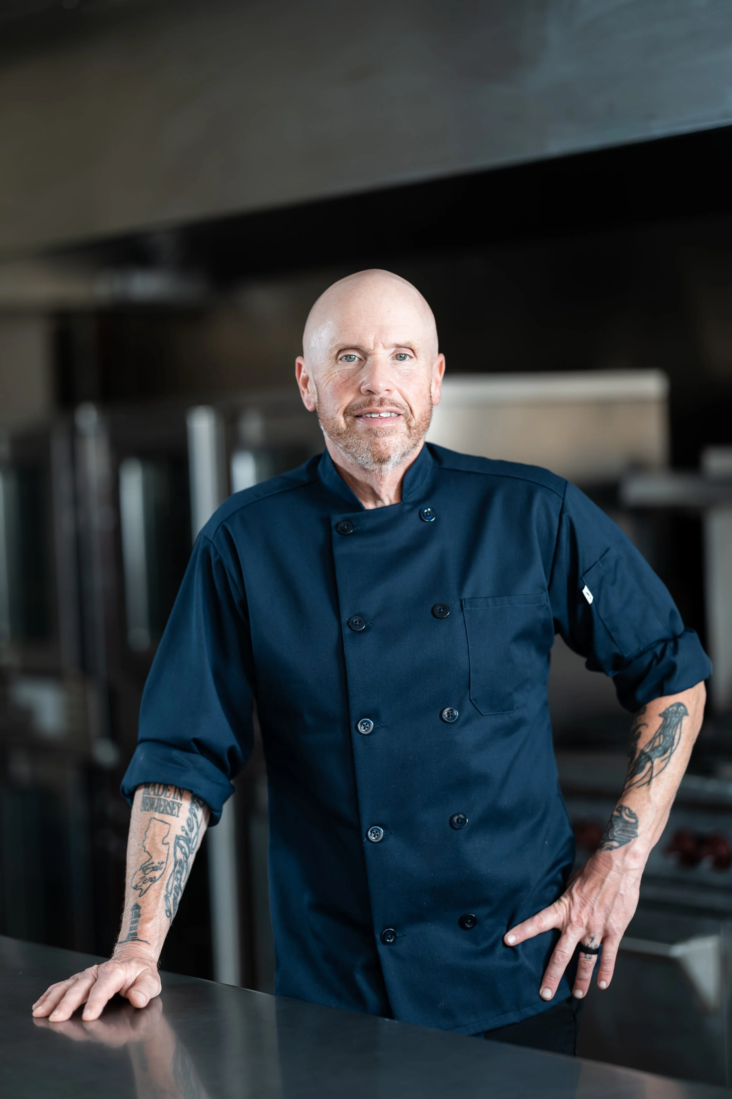 A male chef with tattoos on his arms, wearing a navy chef's coat, standing in a commercial kitchen with stainless steel surfaces and appliances.