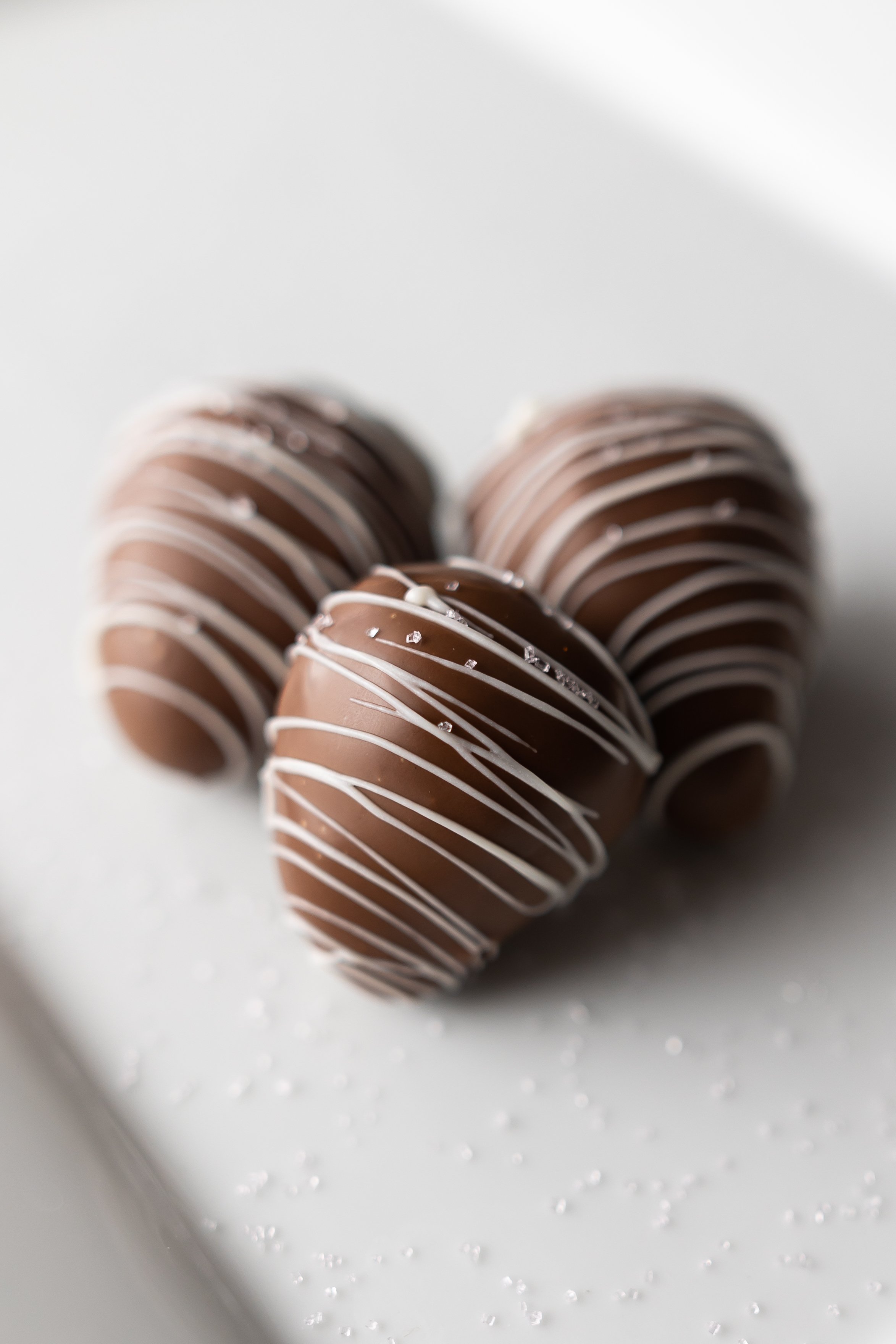 Three chocolate truffles with white chocolate drizzles, sprinkled with coarse sugar on a white surface.