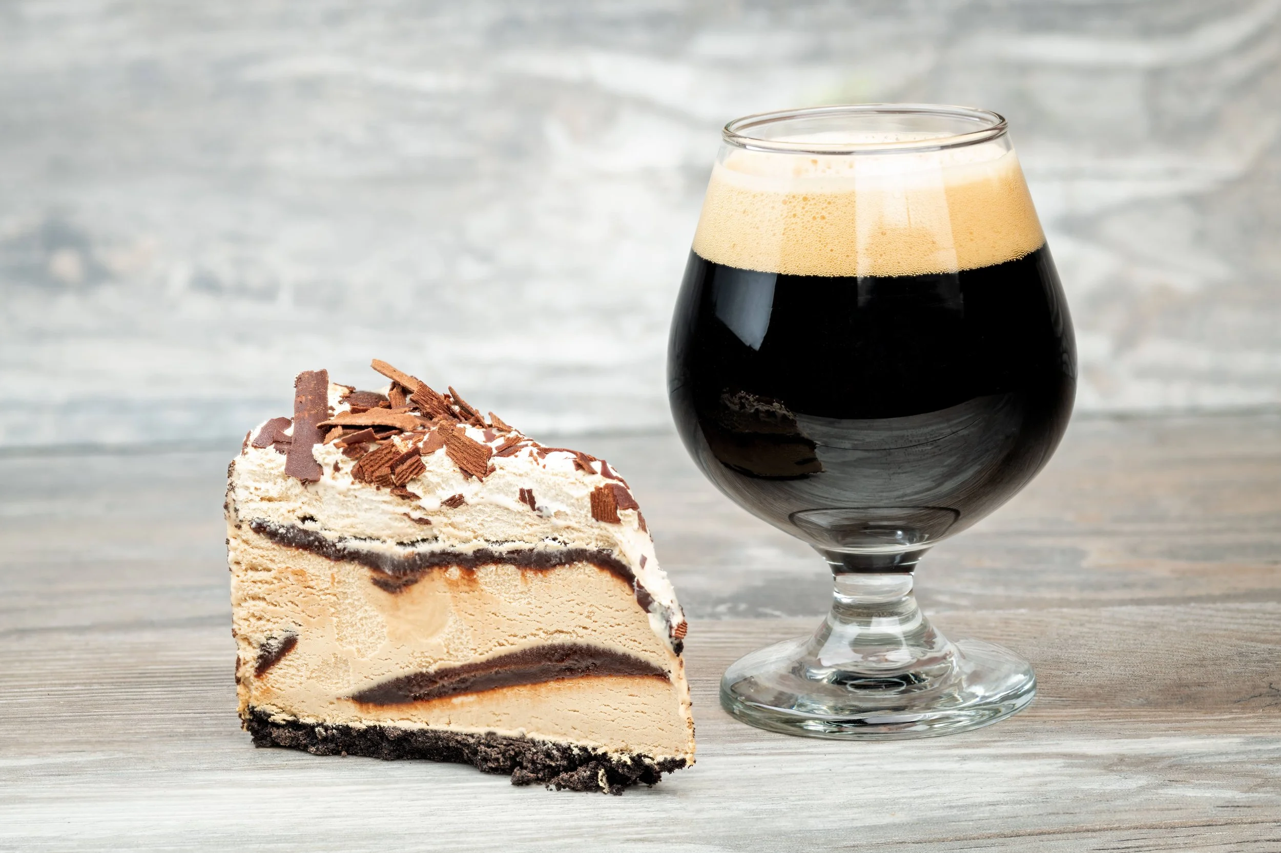 A slice of cookies and cream cheesecake with chocolate shavings next to a glass of stout beer on a wooden surface.