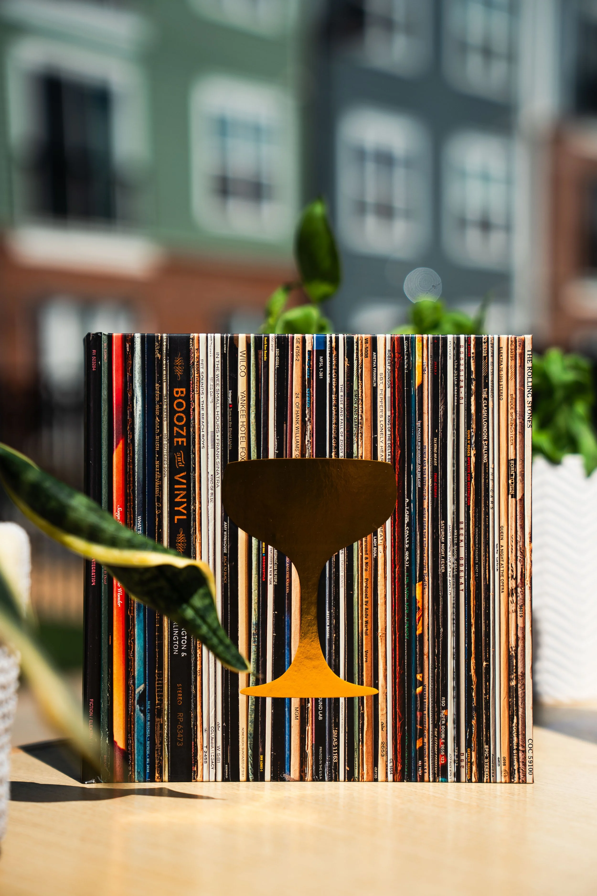 A decorative book featuring a gold-colored coupe glass silhouette in front of a backdrop of colorful vinyl record spines, with some green plants on the sides atop a wooden mobile bar