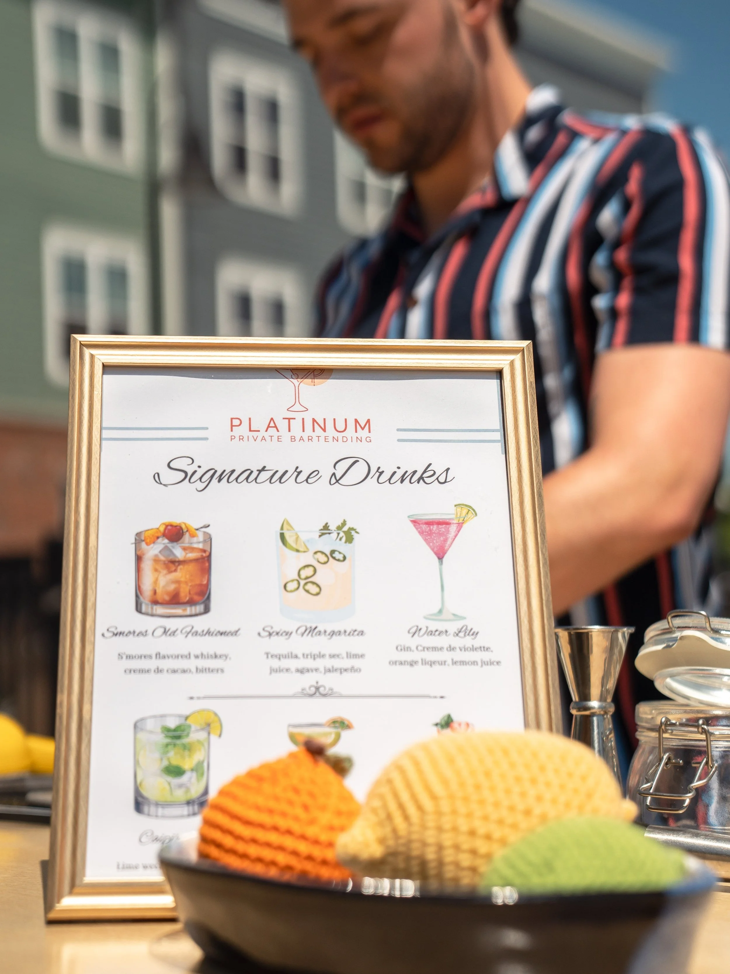 Close-up of a menu from Platinum Private Bartending with signature drinks, featuring colorful illustrations of cocktails, and a bowl of knitted decorative balls in orange, yellow, and green in the foreground. A man wearing a striped shirt is blurred in the background outdoors.