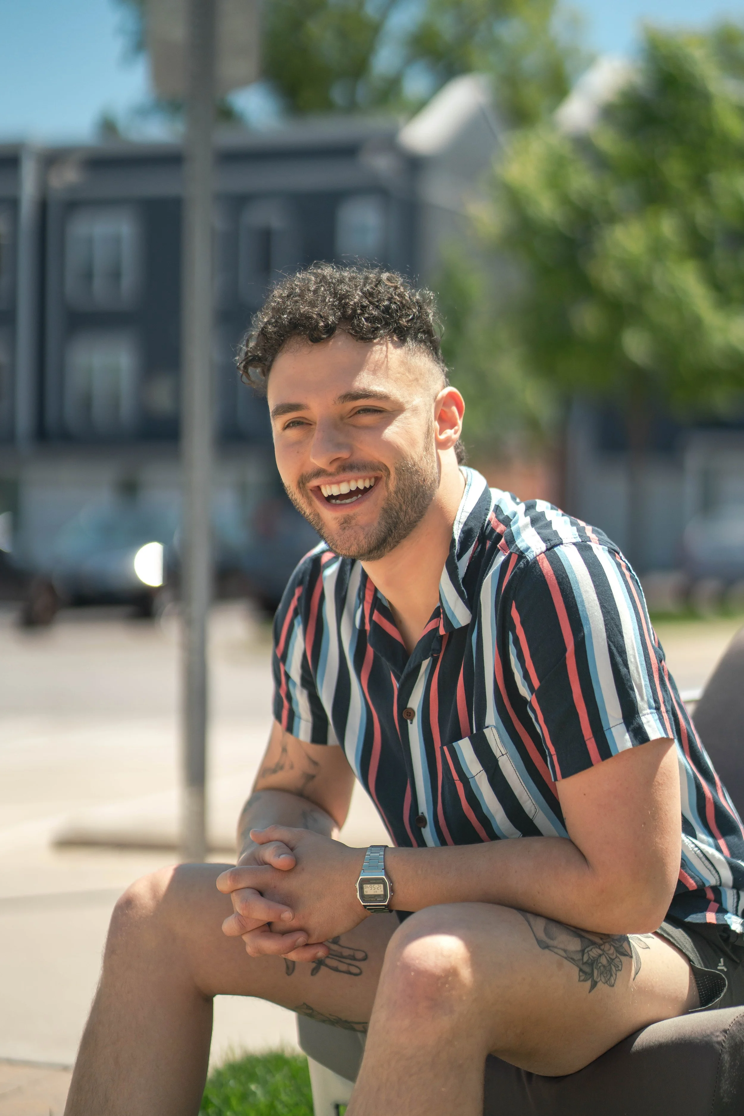 Smiling man with short curly hair, a beard, Tattoos on his legs, wearing a striped shirt and a watch, sitting outdoors on a sunny day.