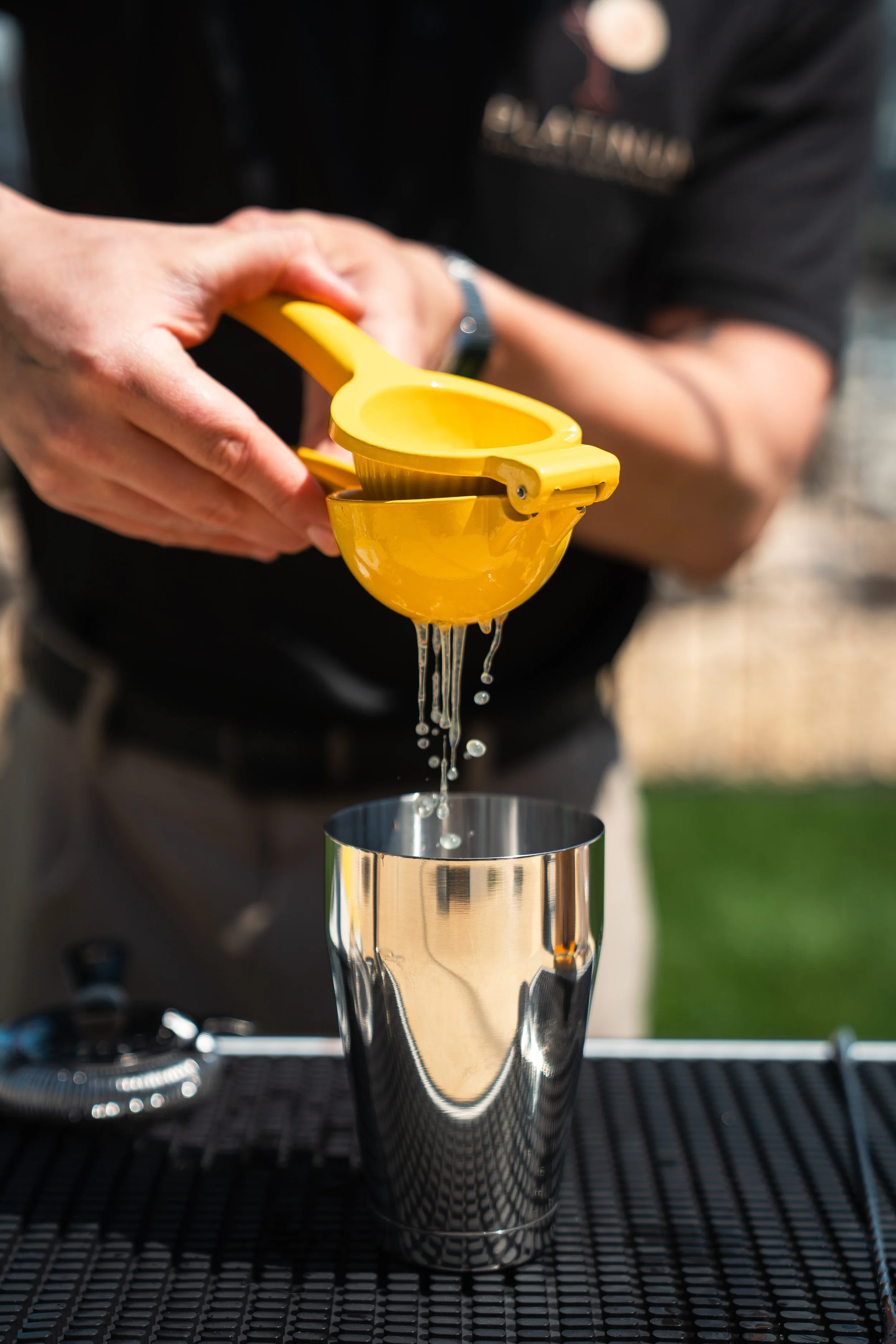 A person is pouring lemon juice through a yellow citrus squeezer into a stainless steel cocktail shaker on a black rubber bar mat.