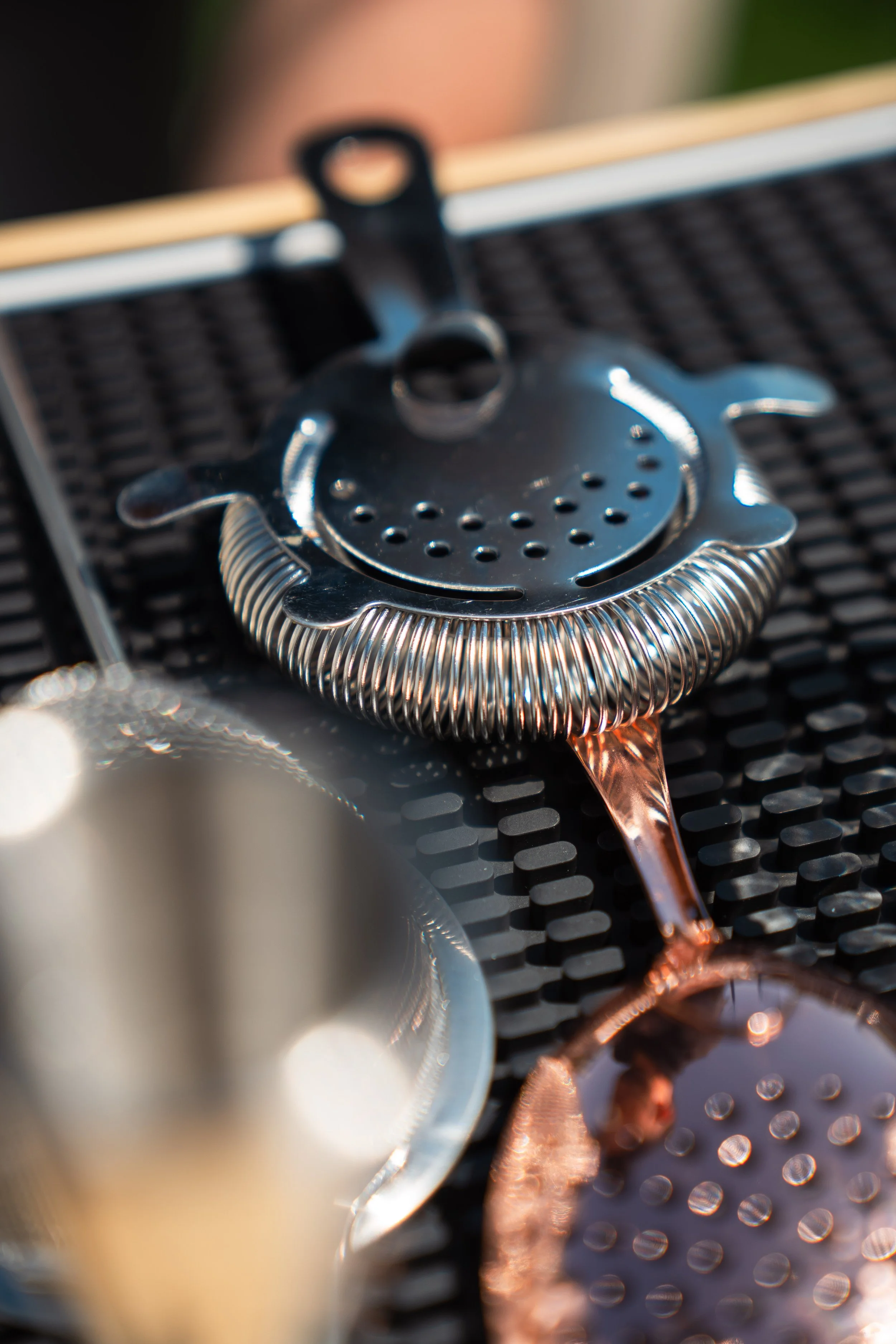 A close-up view of a metallic cocktail strainer resting on a bar mat with a copper-colored cocktail spoon nearby and a blurred drink in the foreground.