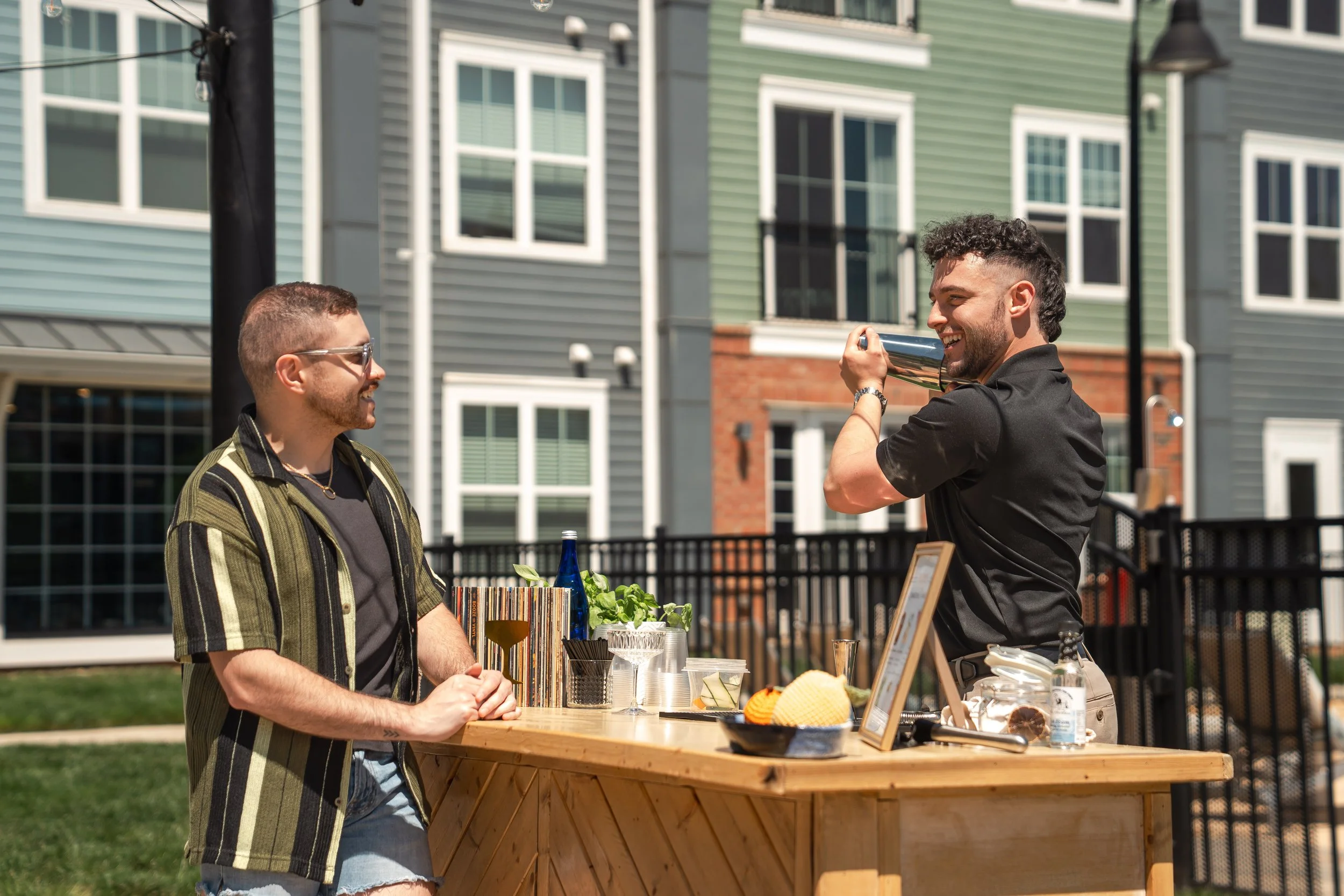 Two men at an outdoor bar; one is bartender shaking a cocktail shaker, the other is customer smiling with hands on the bar, working an event like a wedding or party