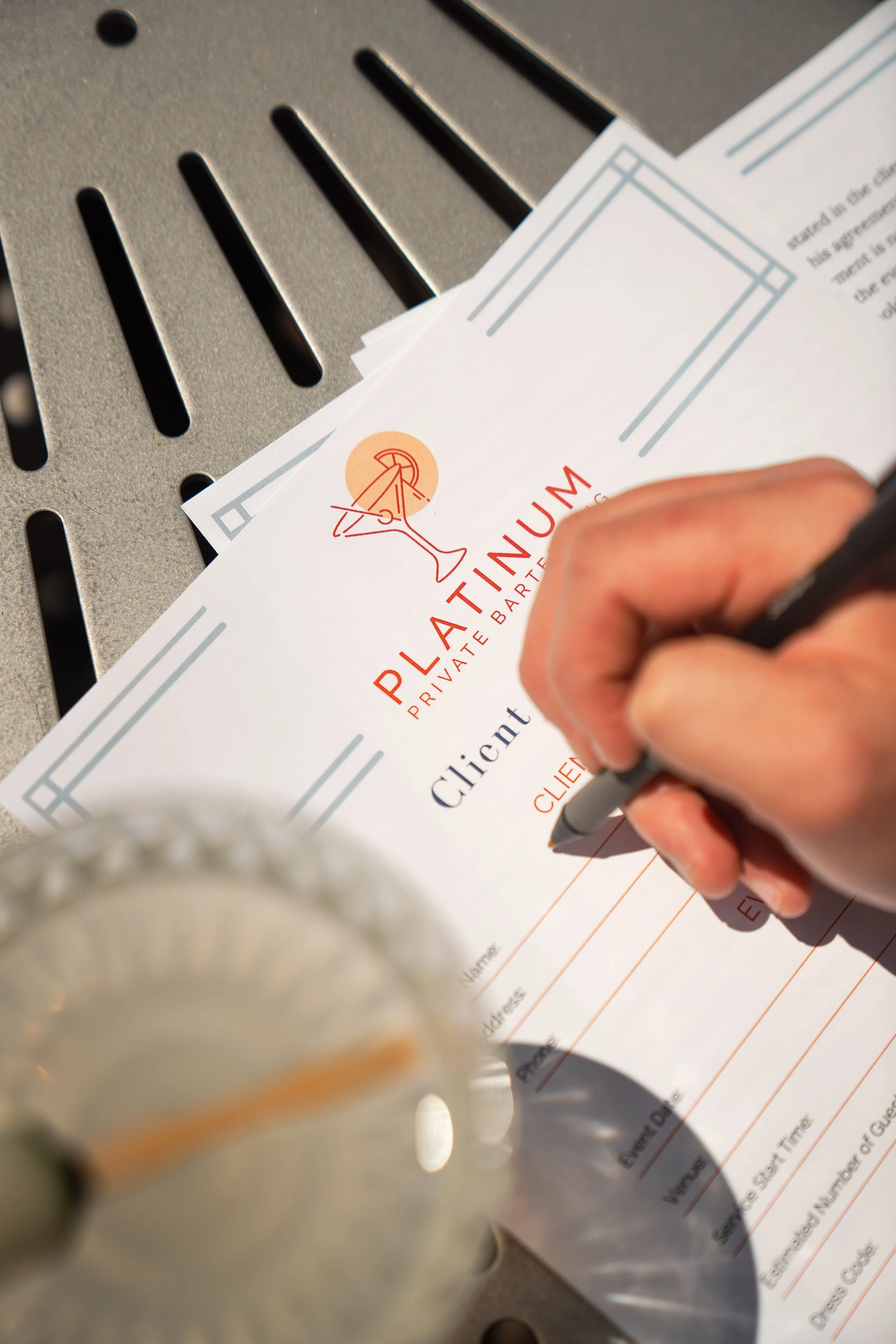 A person filling out a private event booking form for Platinum Private Bartending, on a table with a drink and a black pen.