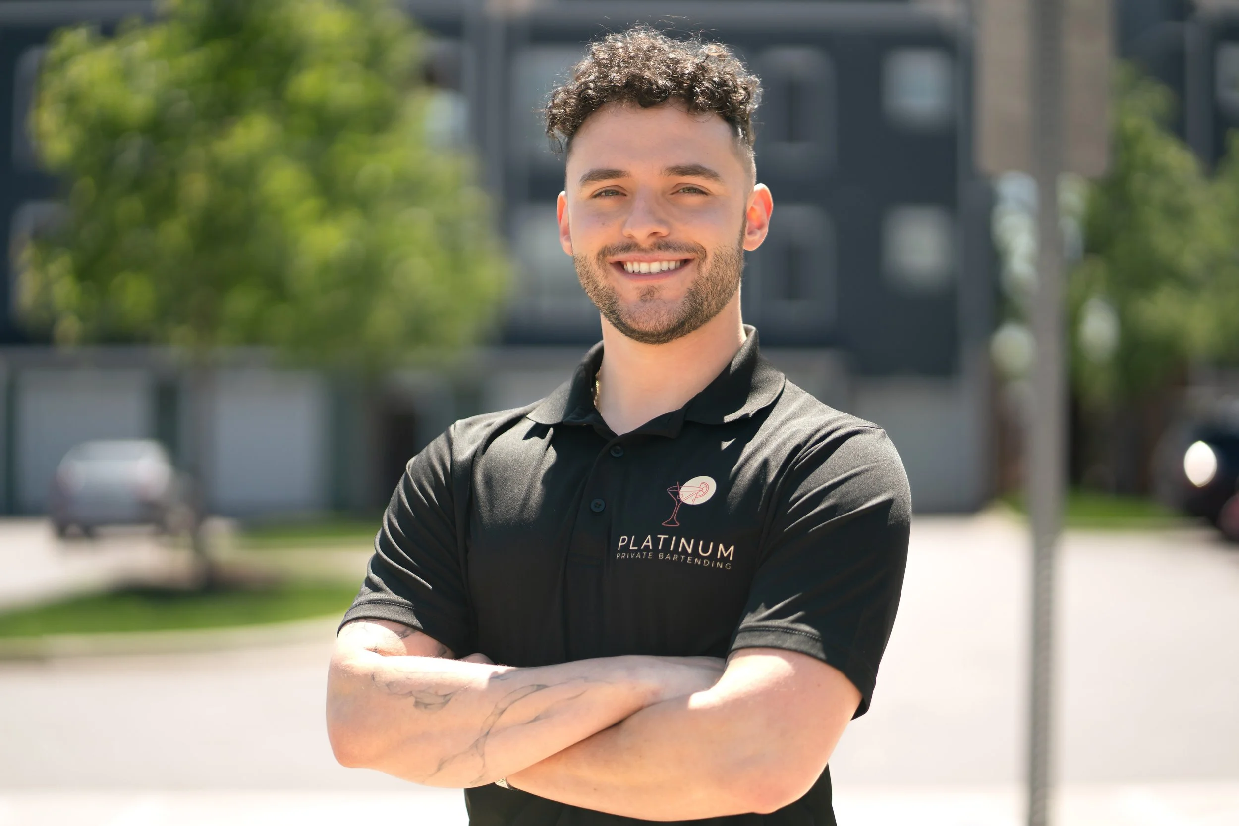 A smiling man with curly hair and a beard, wearing a black polo shirt with the logo 'Platinum Private Bartending,' standing with crossed arms outdoors in front of a blurred background of trees and buildings.