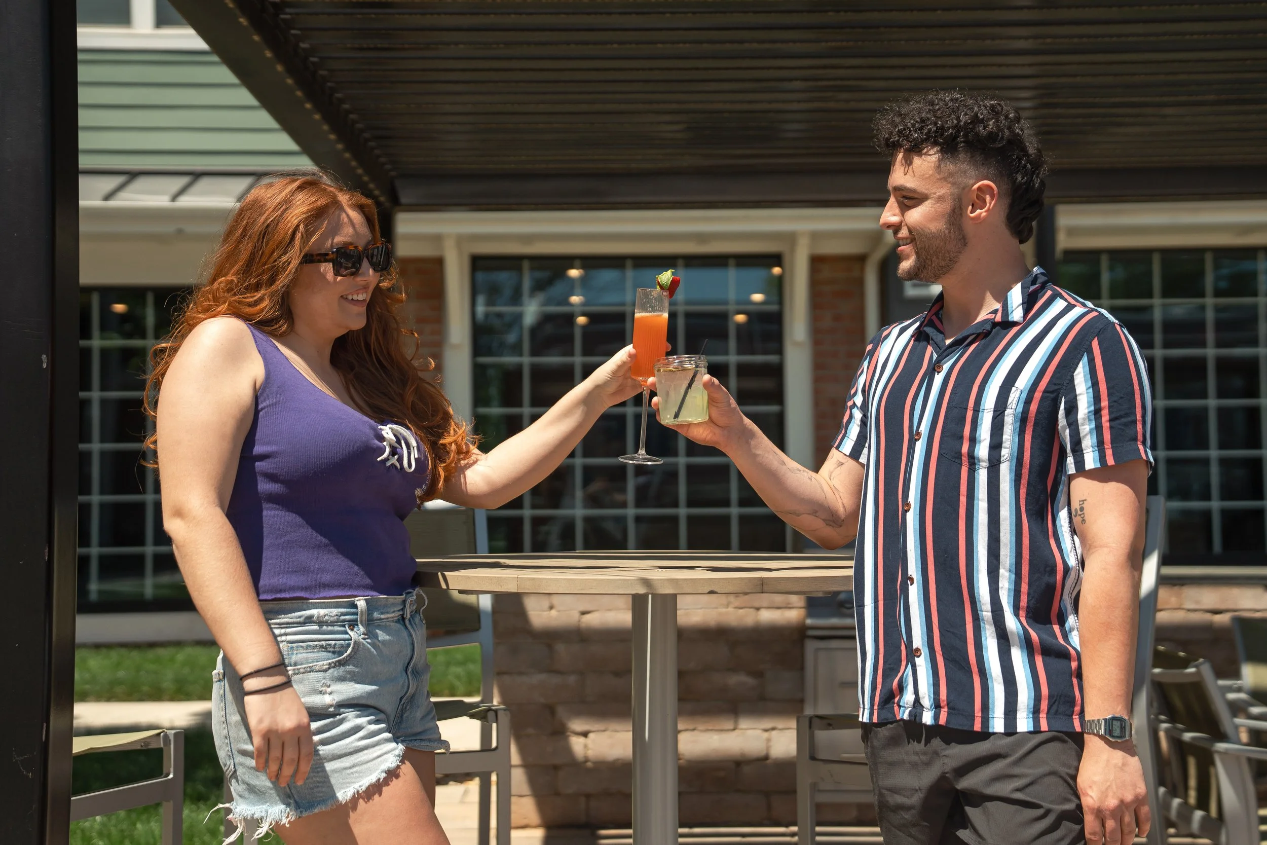 A man and woman are celebrating outdoors on a sunny day, holding cocktails, at a high table . The woman has long red hair, wearing sunglasses, a purple tank top, and denim shorts. The man has dark curly hair, wearing a striped short-sleeve shirt and black short