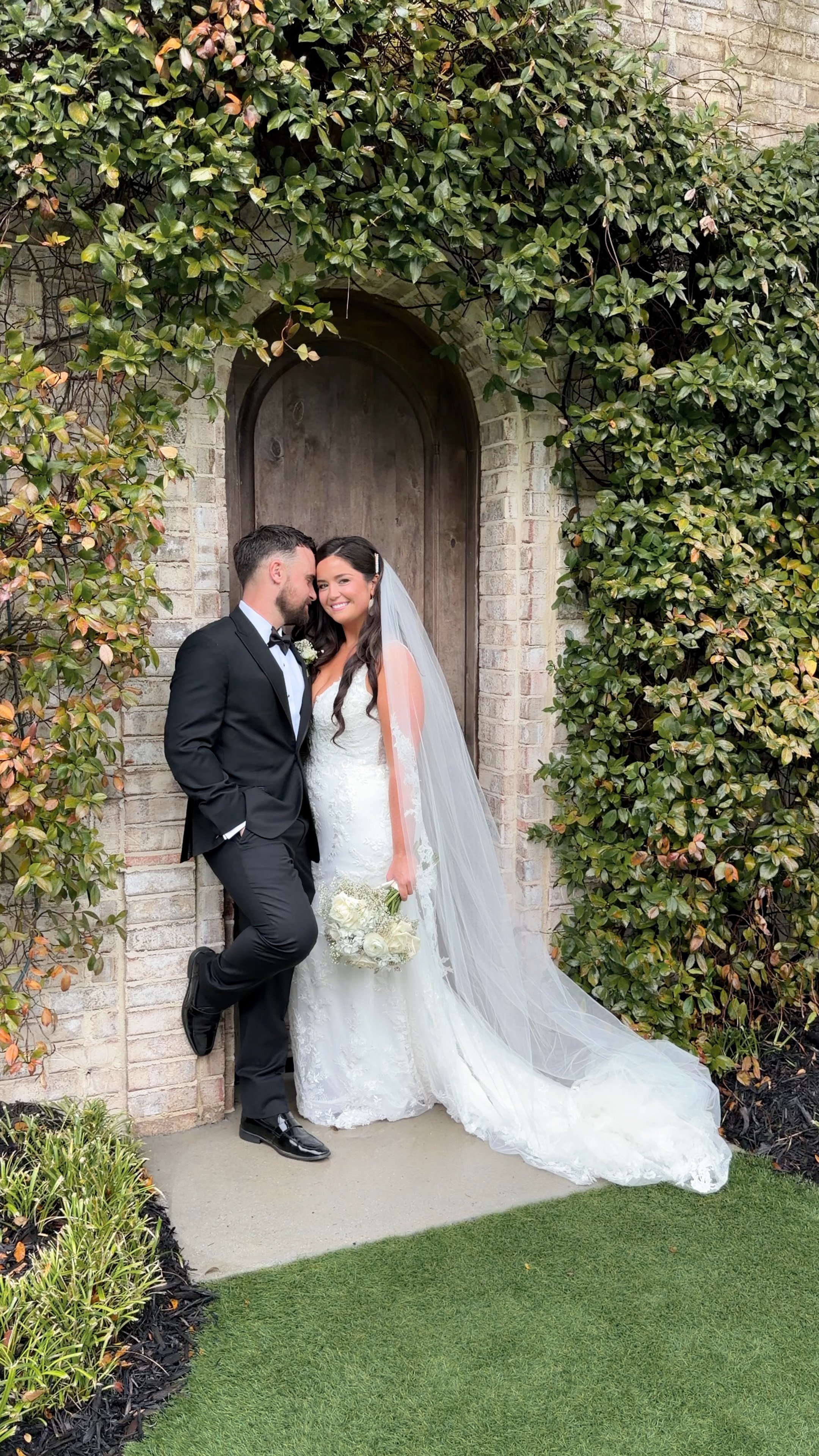 A bride and groom in wedding attire standing by a brick wall with greenery at wrightsville manor by wilmington content co - content creator in north carolina