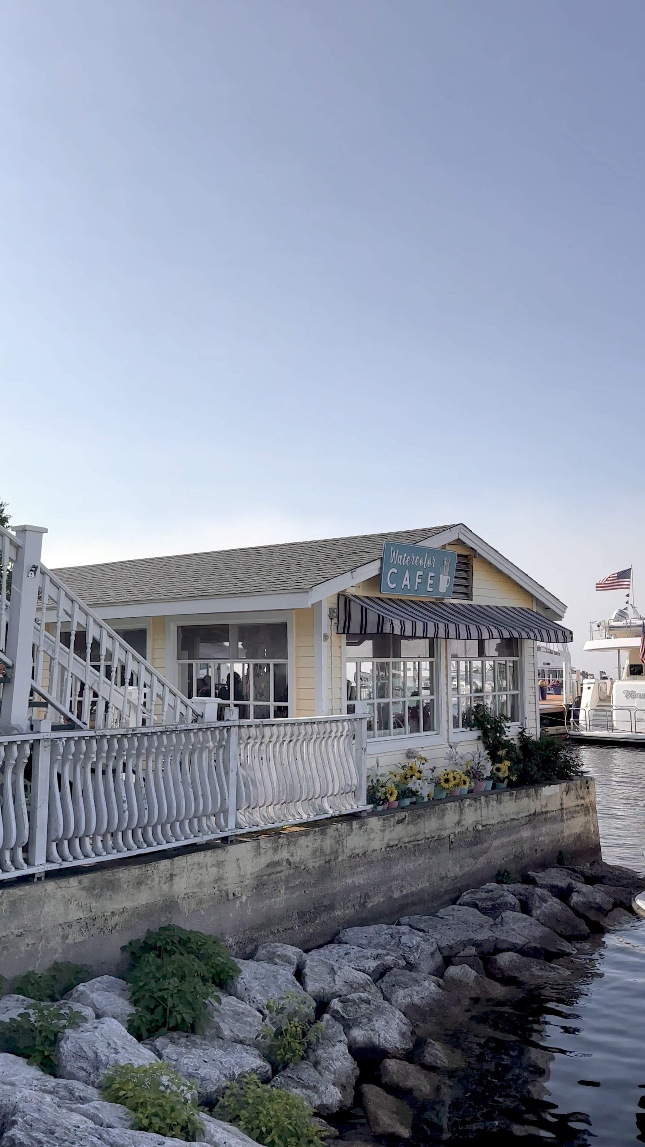 A seaside cafe named 'Watercolor Cafe' with a yellow exterior, white railing, and striped awning, situated by a rocky shoreline with boats in the background and an American flag flying.