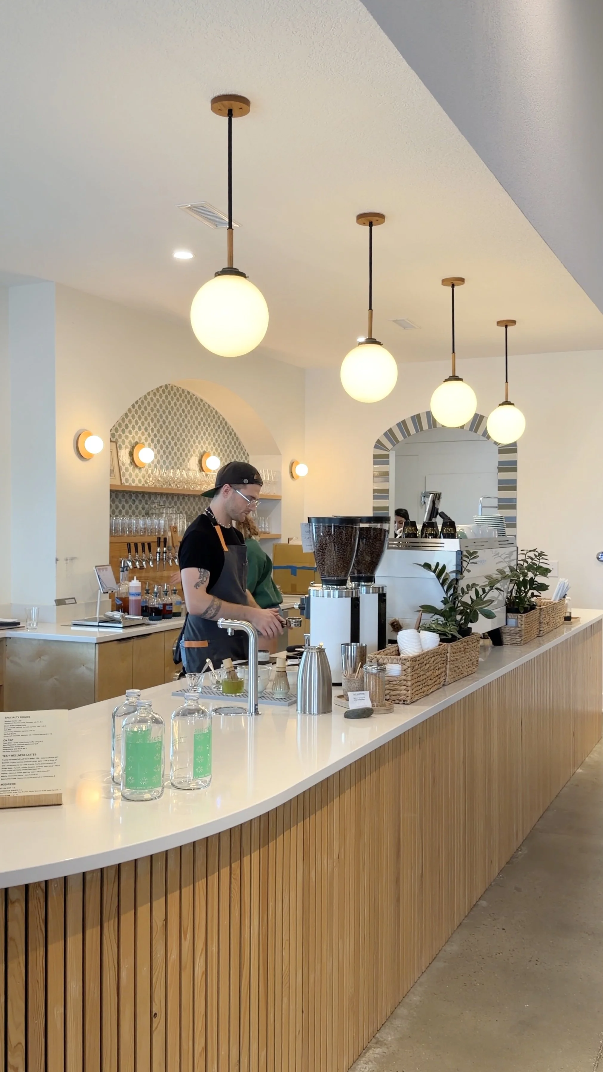 Inside a modern coffee shop with warm lighting, wooden accents, and pendant lamps hanging from the ceiling, a barista prepares drinks behind the counter with shelves of glassware and coffee equipment.