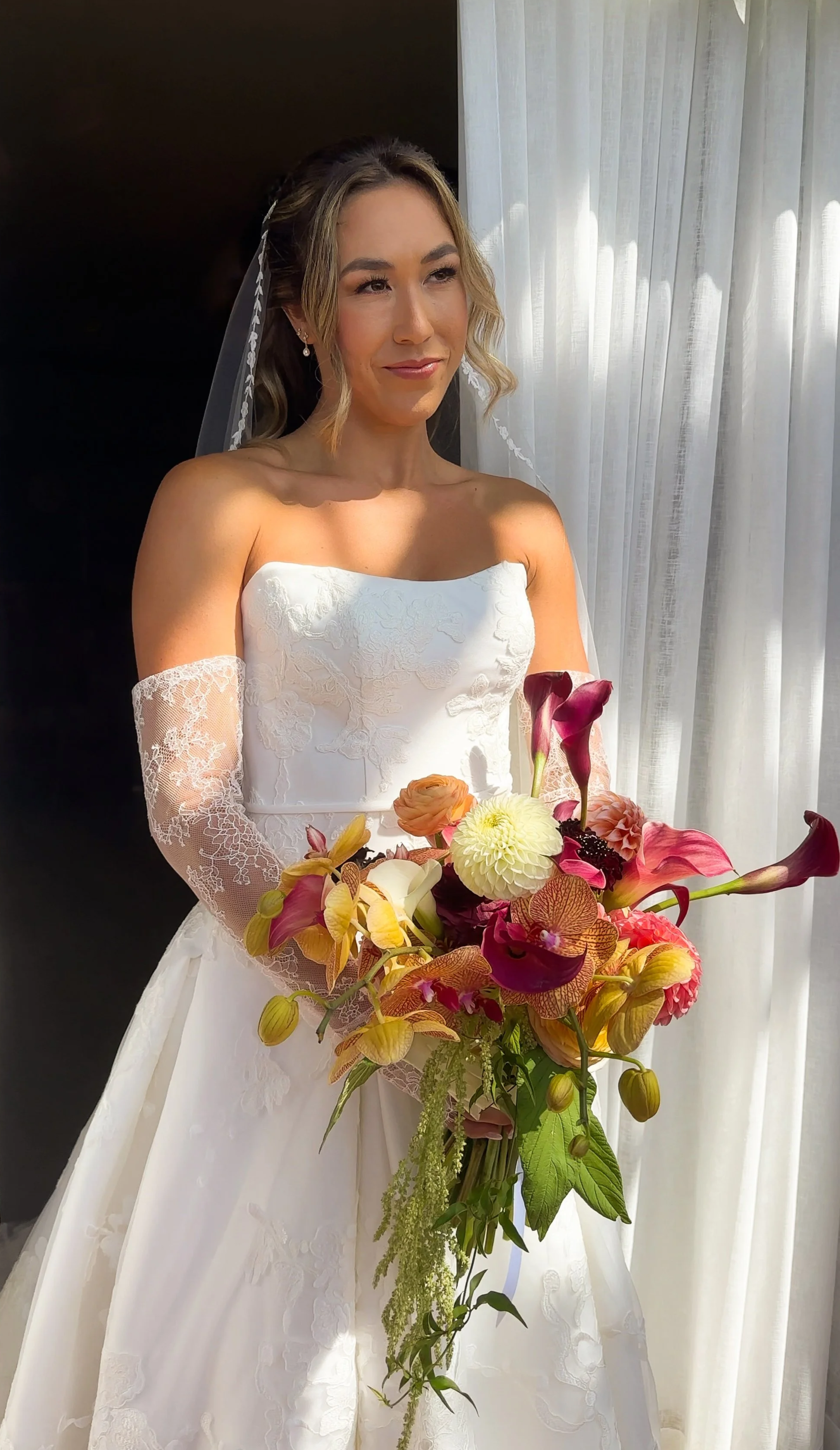 A bride in a white dress holding a large bouquet of colorful flowers, standing near a white curtain, with sunlight illuminating her face. by wilmington content co - content creator in north carolina