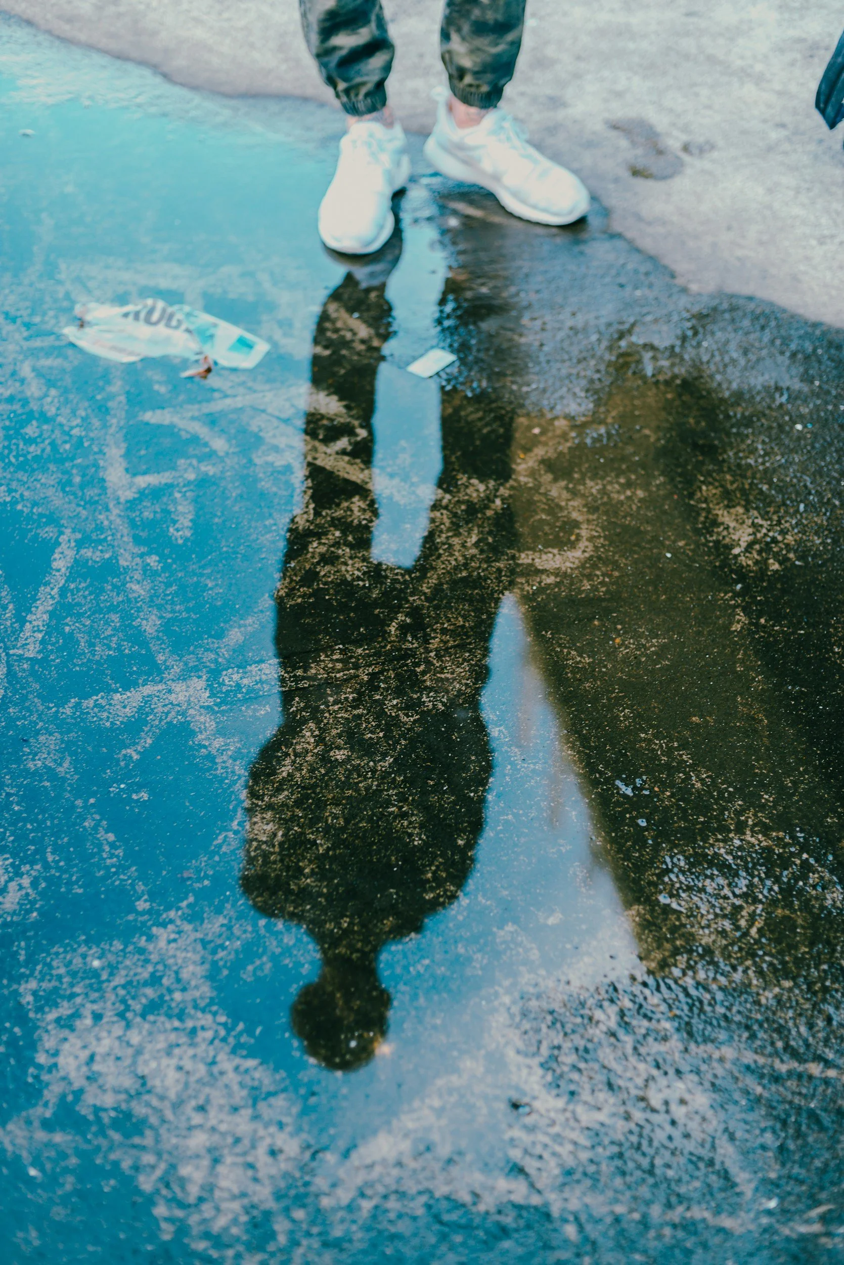 Reflection of a person wearing white shoes and dark pants in a puddle on a wet surface, with some trash nearby.
