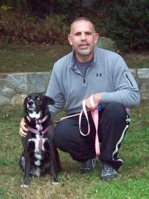 A man crouching outdoors next to a black and white dog on a pink leash, with a stone wall and trees in the background.