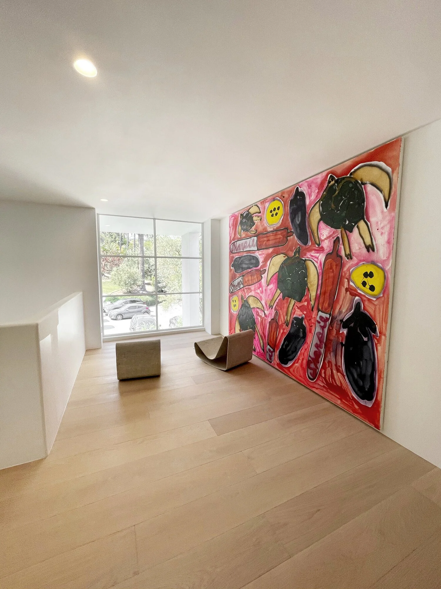 A modern interior room with light wooden flooring, a large window showing a parking lot and trees outside, two small brown chairs, and a large colorful painting of dogs playing with popsicles on the wall.