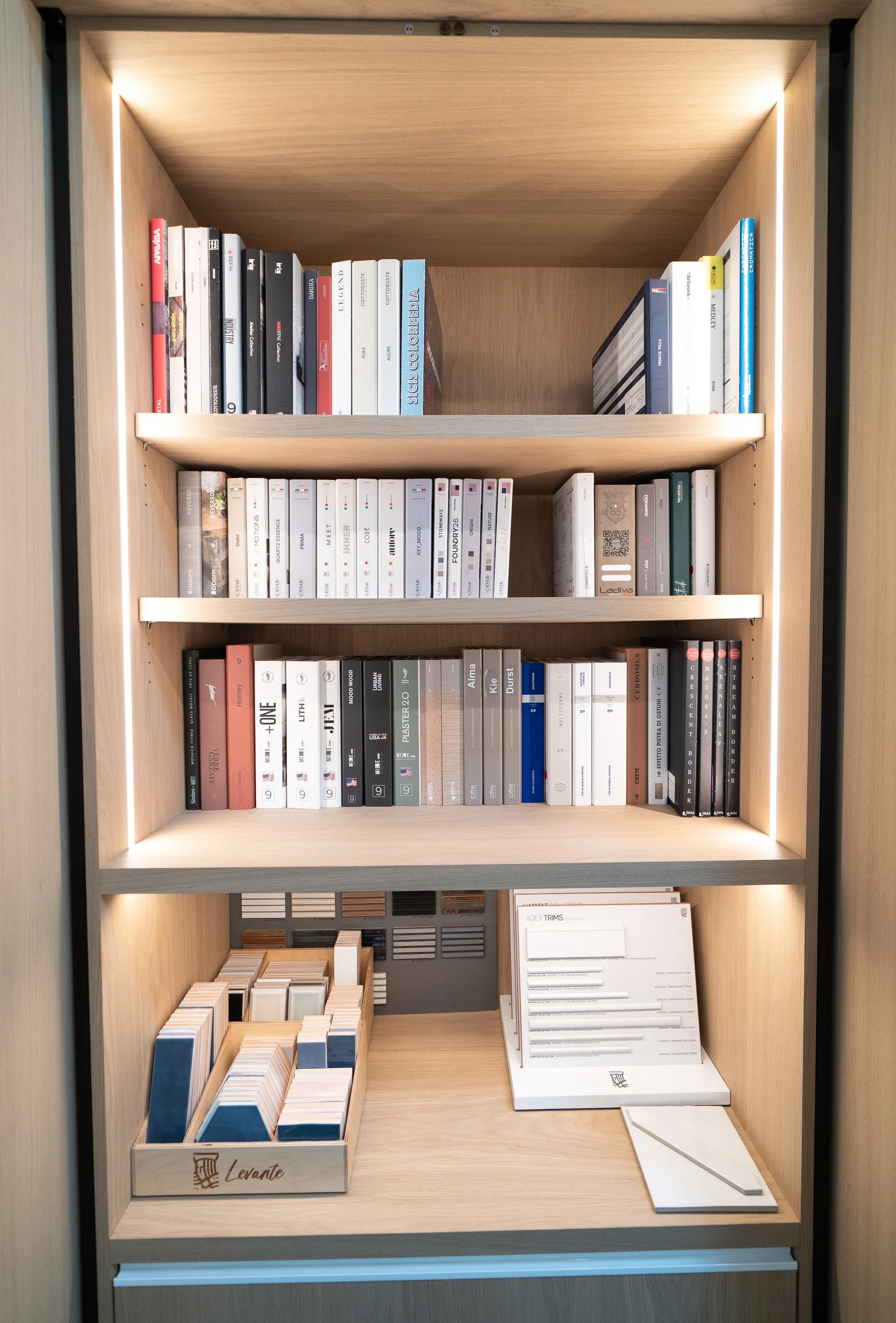A wooden bookshelf with four shelves containing books and samples of material swatches, with lighting accents on the sides and bottom shelf.