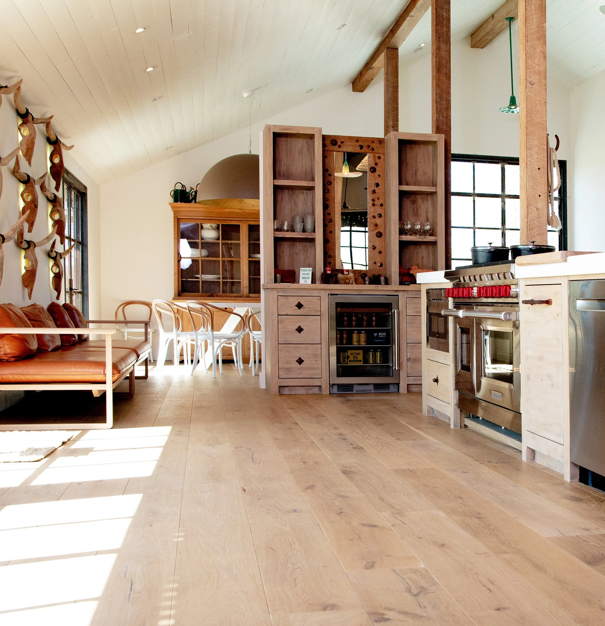 Open-concept kitchen and dining area with wooden flooring, a leather sofa, white chairs, and wooden cabinetry and shelving, decorated with modern and rustic elements.