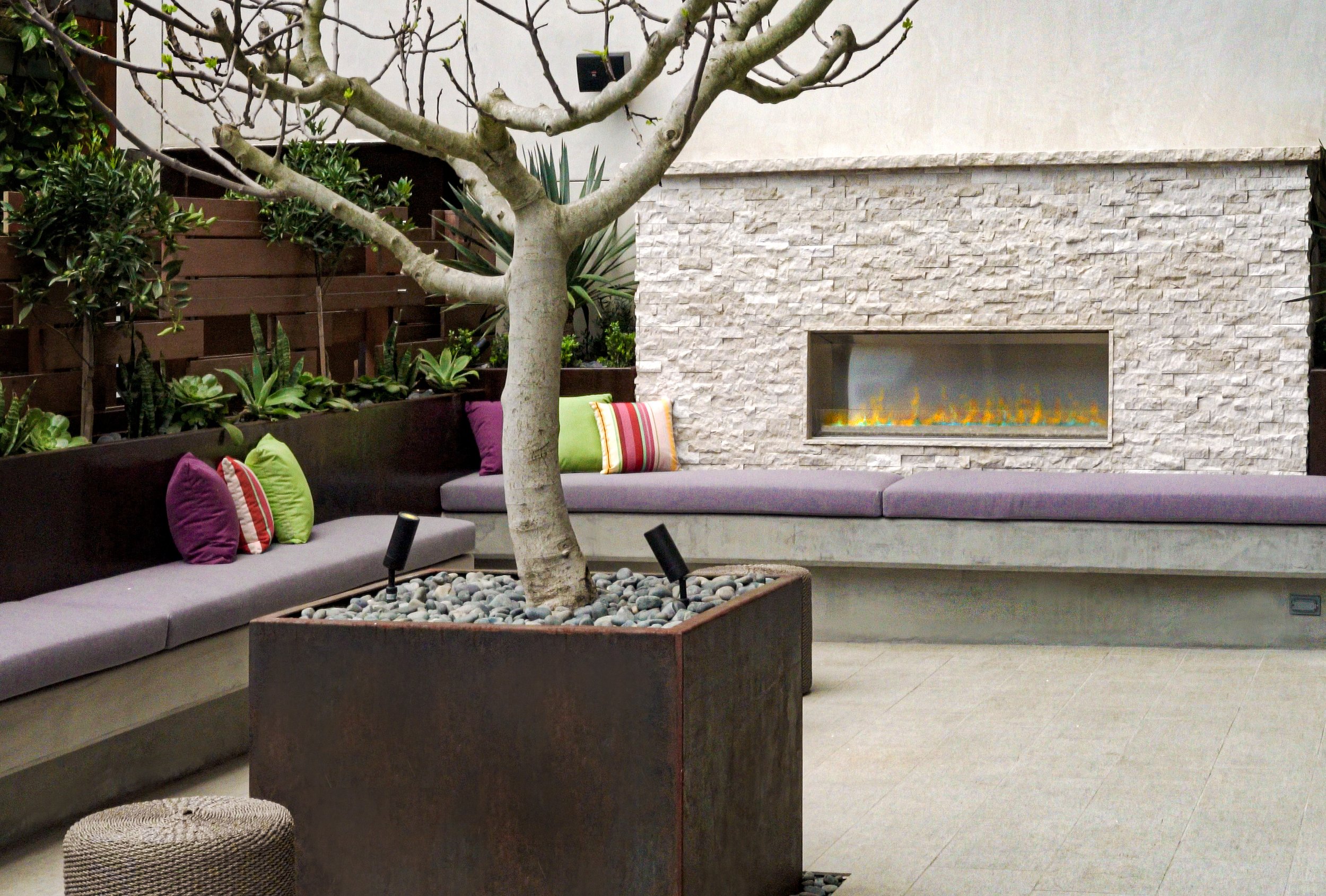 Indoor lounge area with a leafless tree, colorful pillows on built-in gray seating, a white stone fireplace with a small fire, and potted plants along the wall.