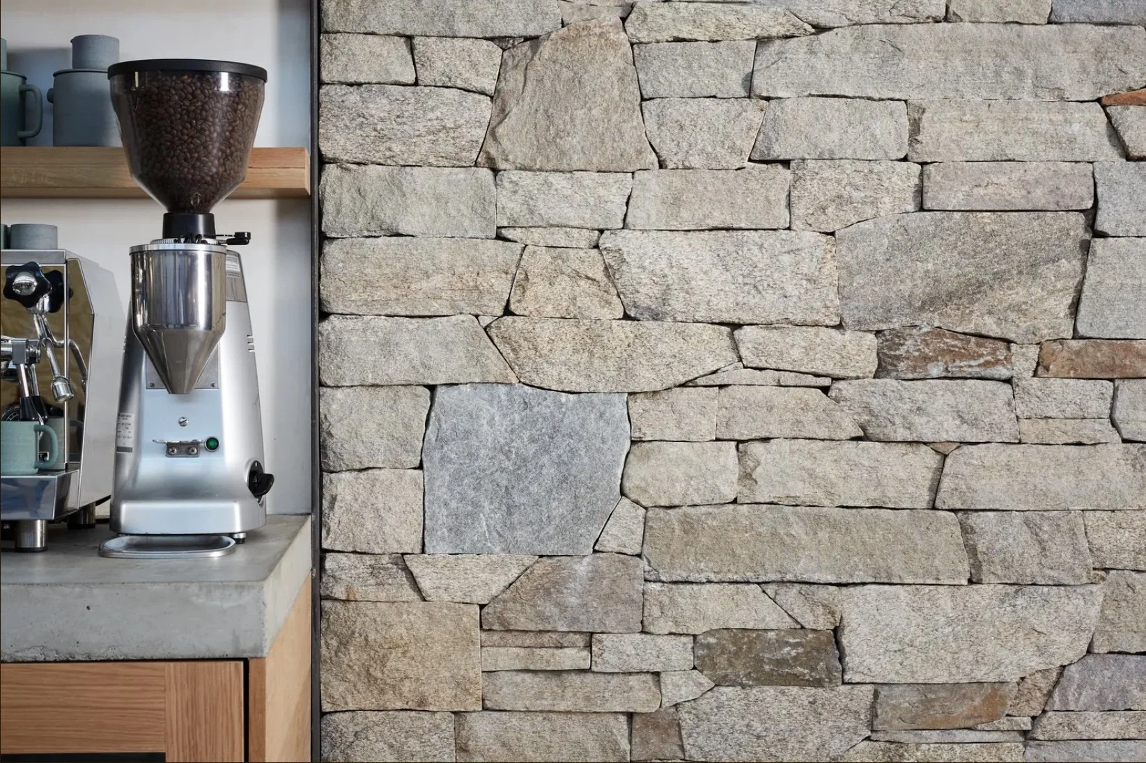 A modern wooden house situated on a hillside with tall grasses, overlooking a large body of water and mountain range in the distance during dawn or dusk.