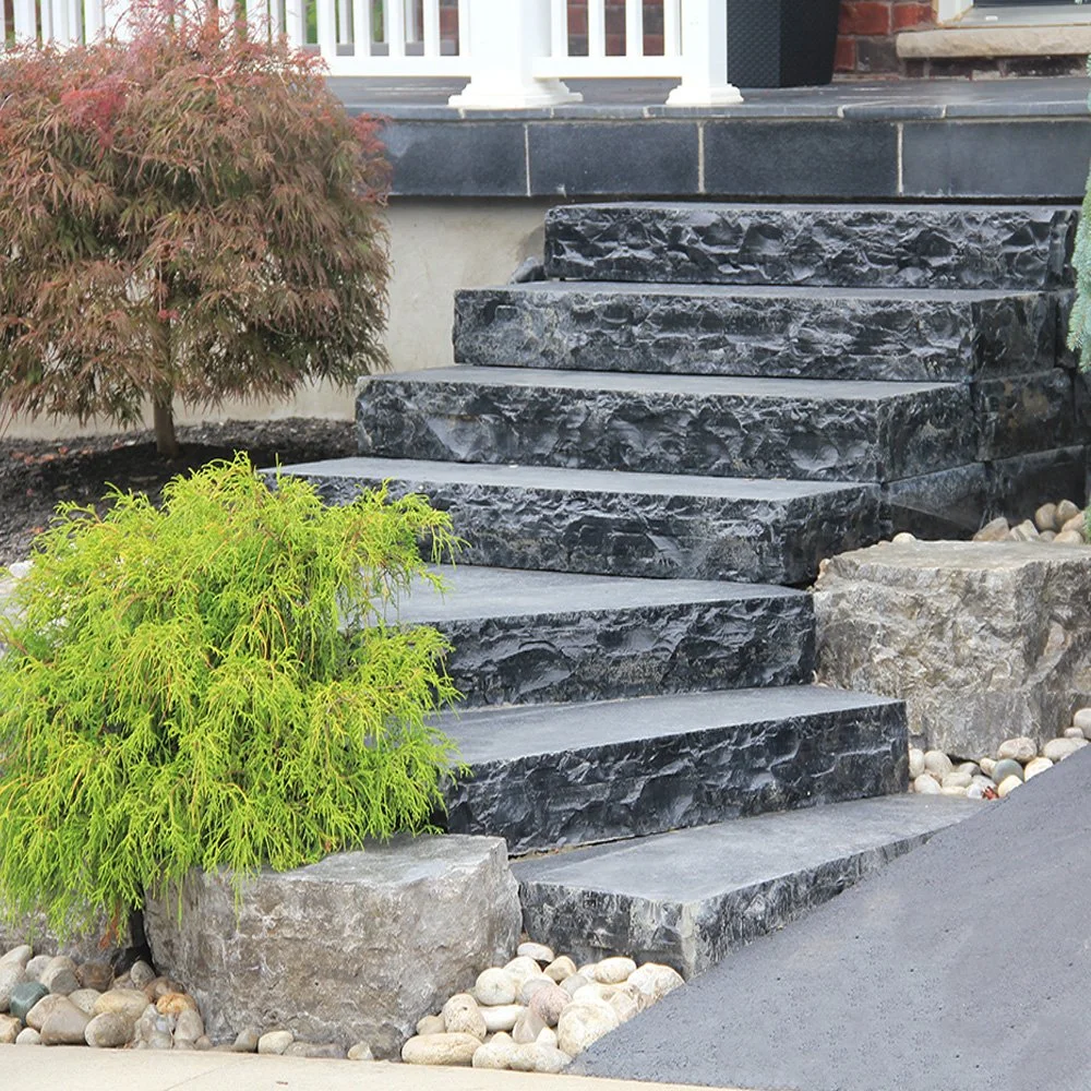 Black stone steps leading up to a porch with a white railing, surrounded by gardening rocks and greenery including a small red-leafed tree and a green shrub.