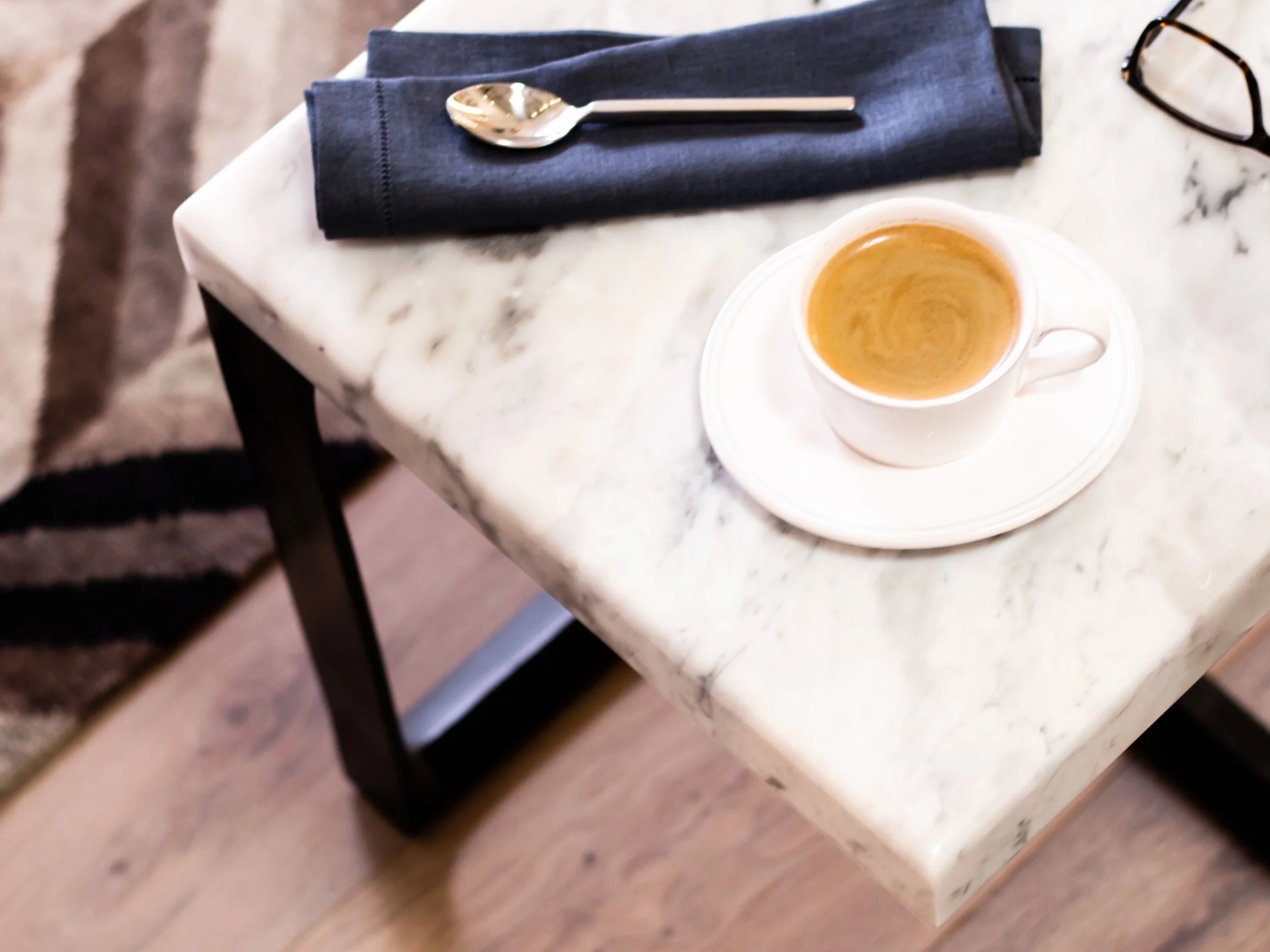 A white coffee cup filled with espresso on a white saucer, placed on a marble table with a dark napkin, spoon, and eyeglasses nearby.