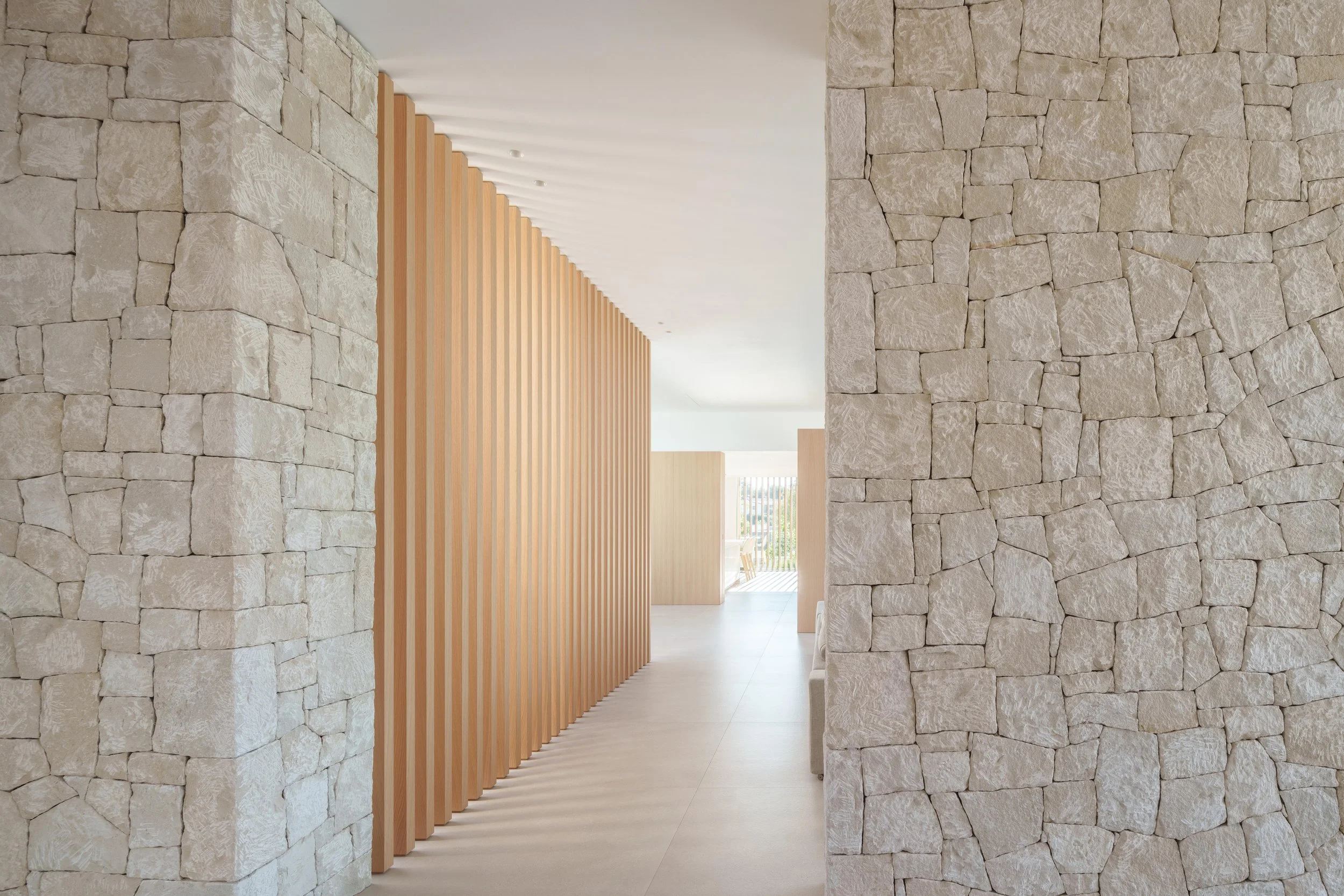 Interior view of a modern building with stone walls, a wooden slat partition, and a bright space with a view of the outdoor area.