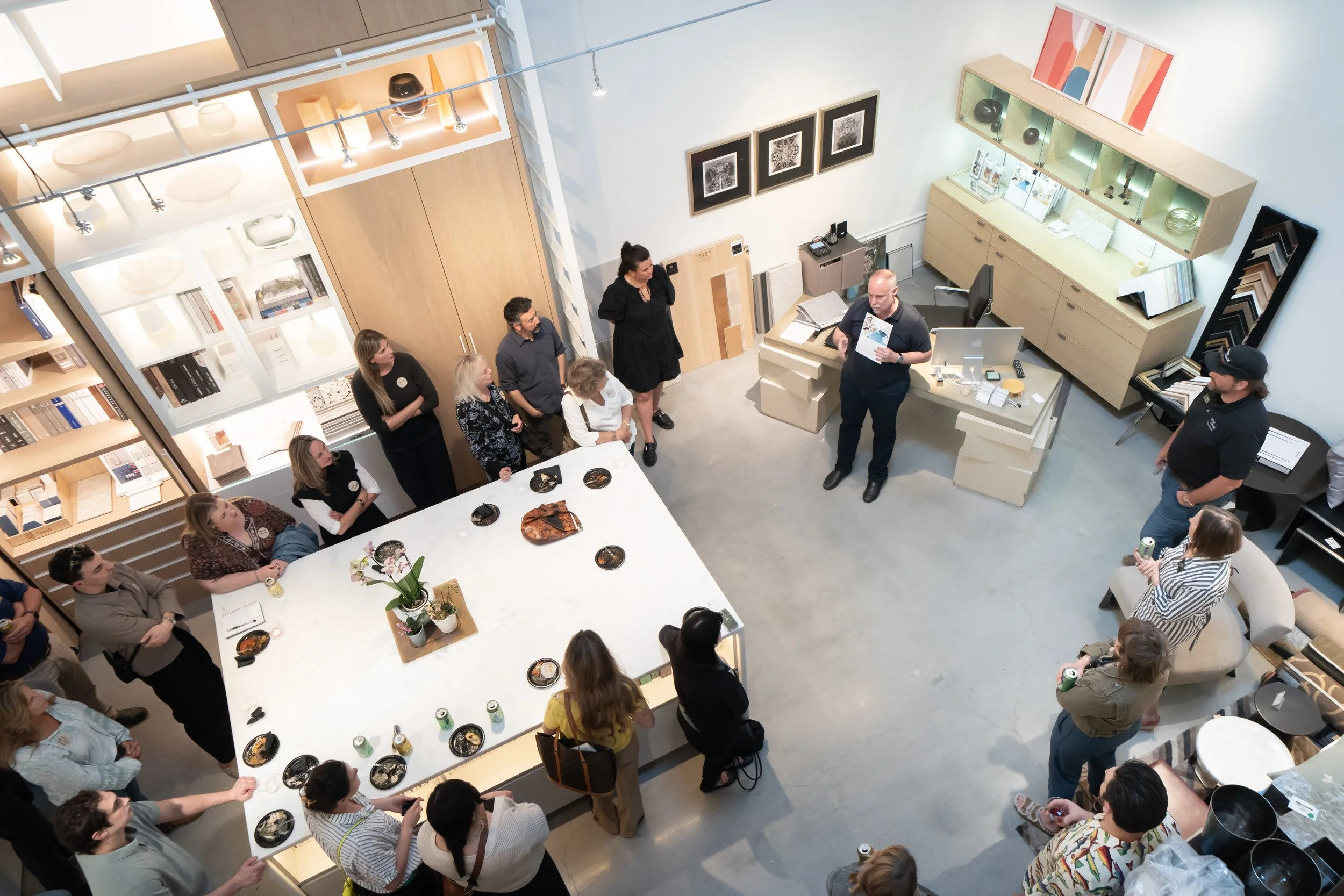 A group of people attending a presentation or class in a modern showroom or gallery, with displays of home decor and furniture around, as one man stands reading from a brochure and others listen.