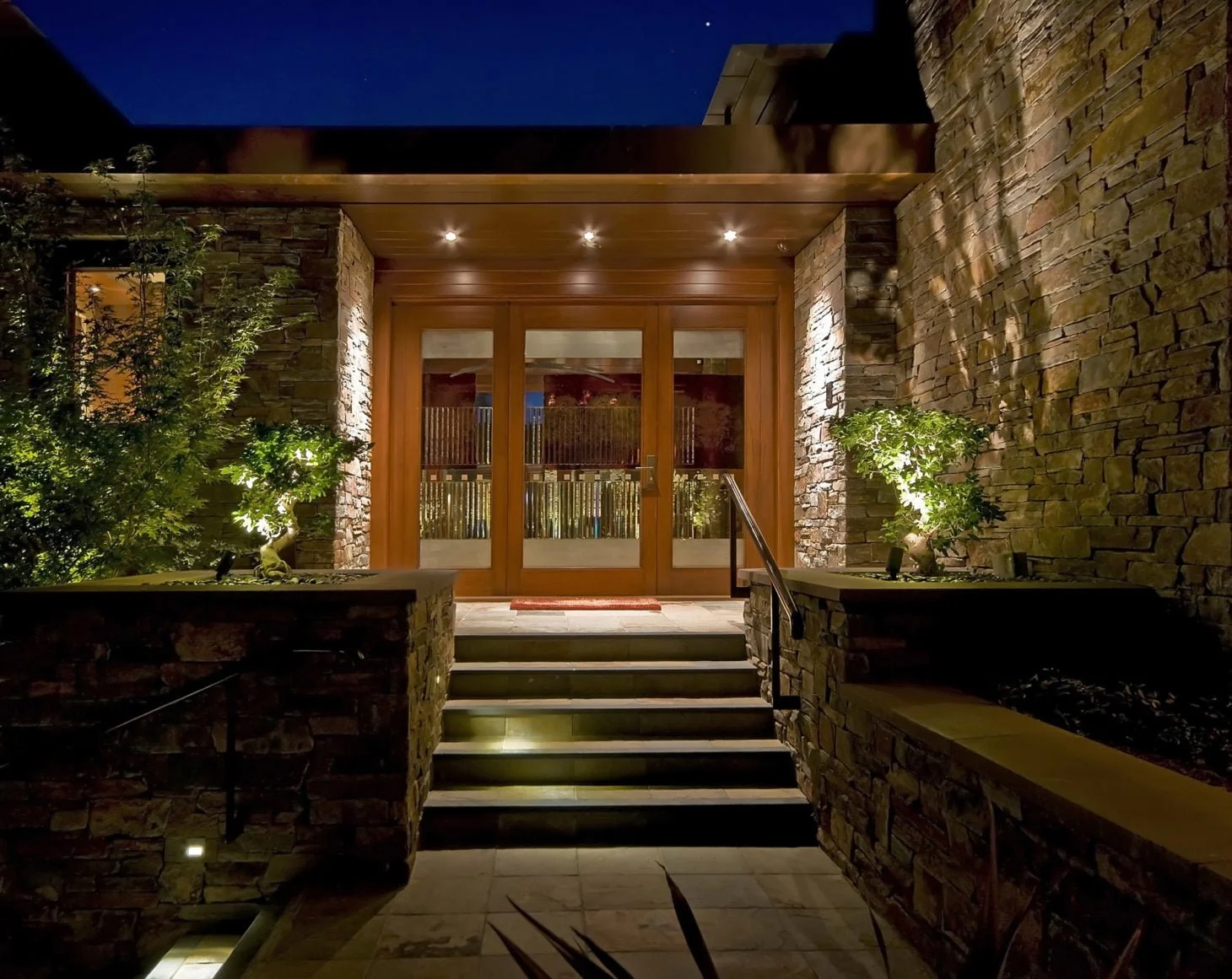 Night view of a home's front entrance with stone steps, wooden door, and exterior lighting highlighting stone walls and small trees.