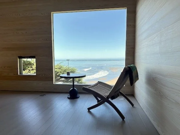 A minimalist room with a large window overlooking the ocean, a lounge chair, and a small round coffee table with a glass of water.