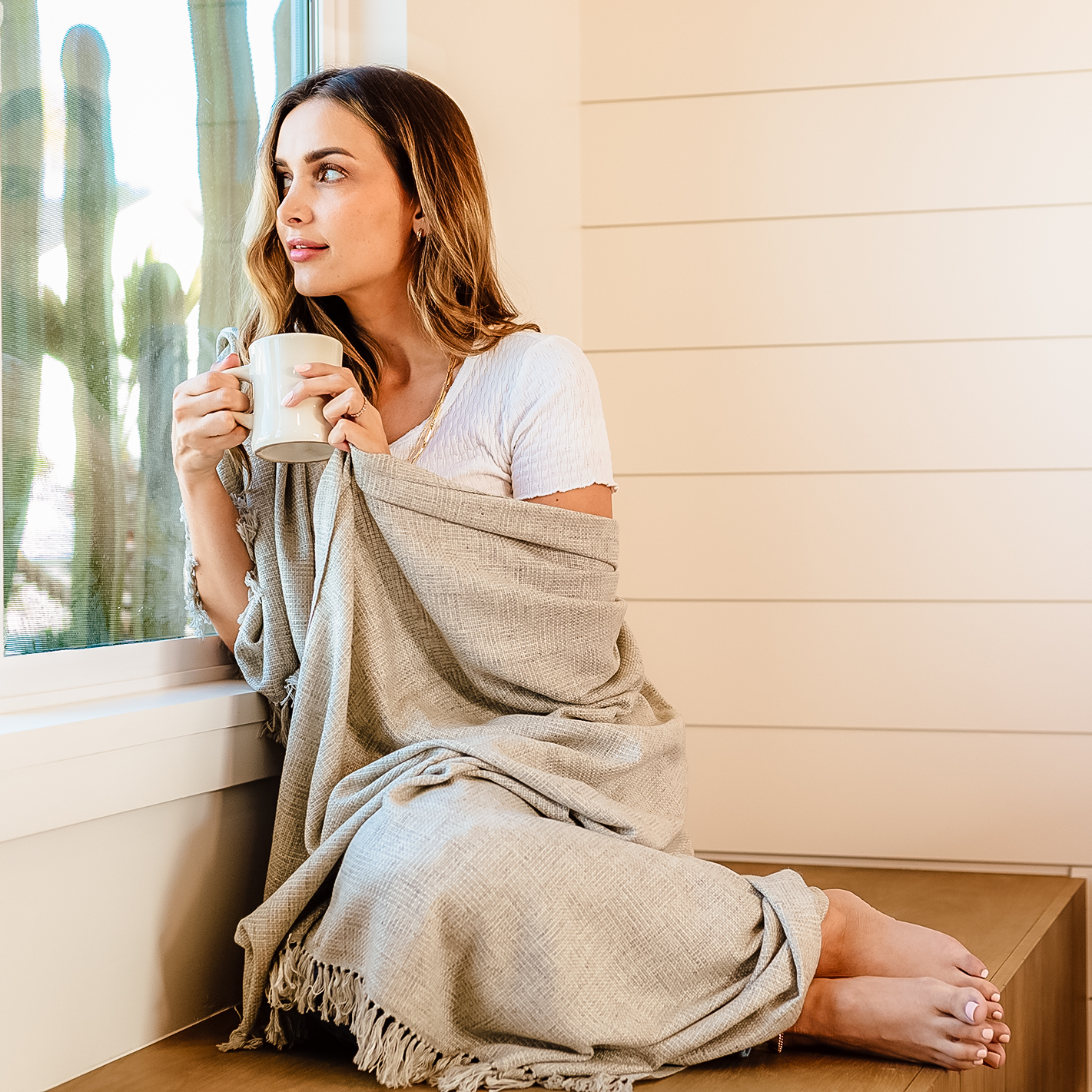 A woman sitting on a window seat holding a white mug and looking outside, wrapped in a beige blanket with a tan wall behind her.