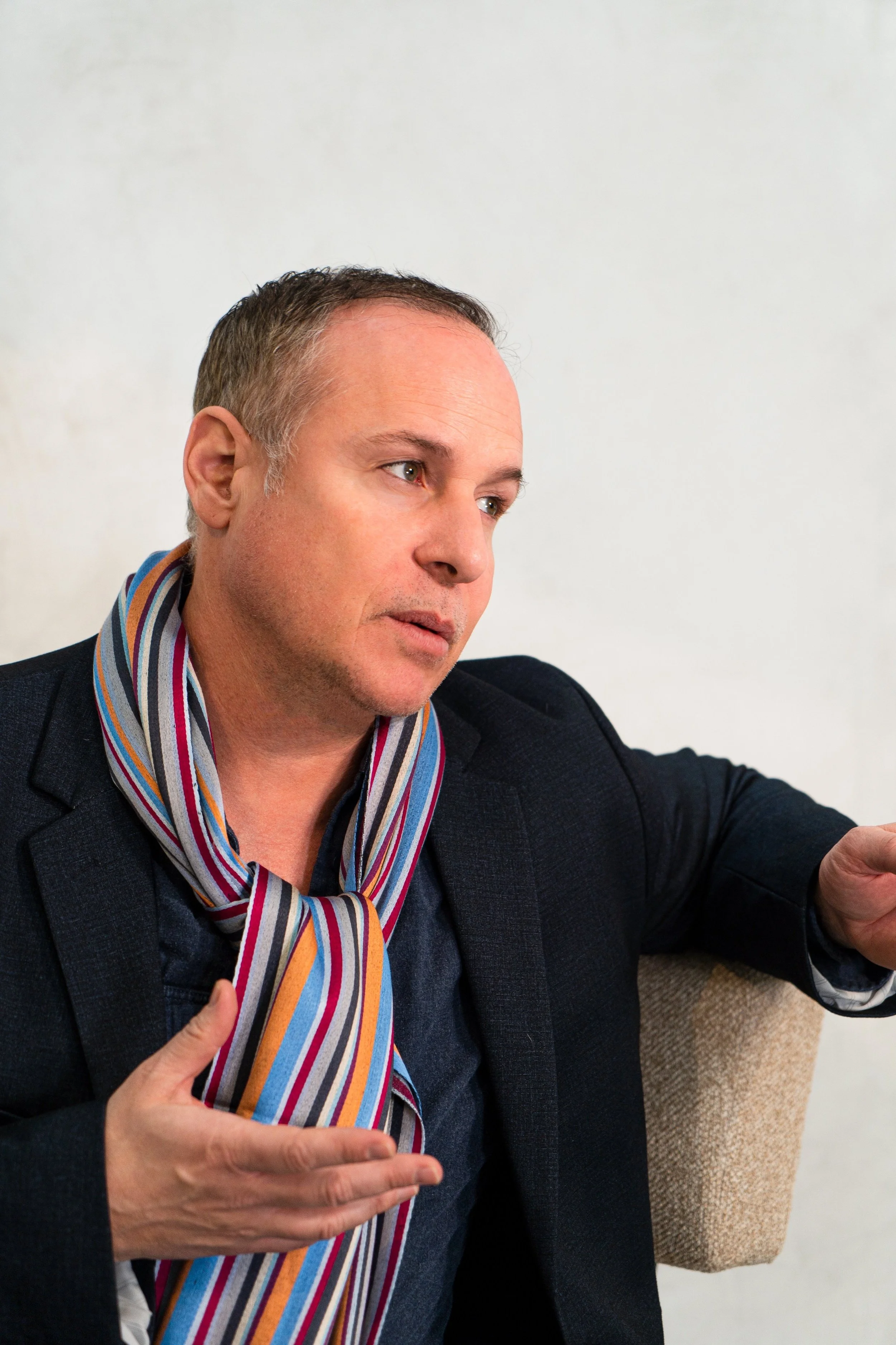 A man with short hair and a serious expression, wearing a dark blazer and a colorful striped scarf, sitting indoors against a plain light-colored wall.