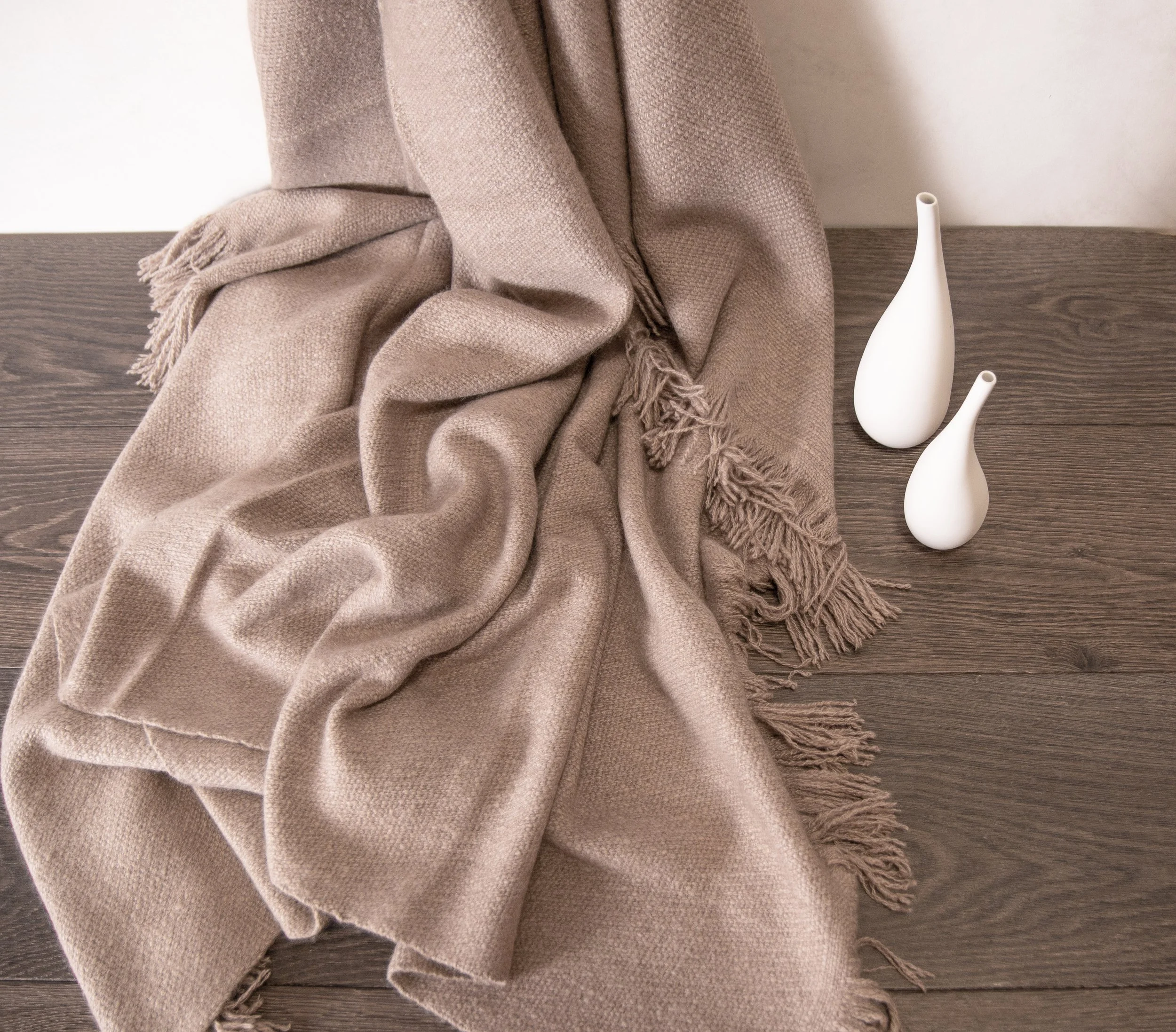 A beige woven throw blanket with fringes draped on a dark wooden surface, next to two white ceramic vases with narrow necks and rounded bases, against a white wall.