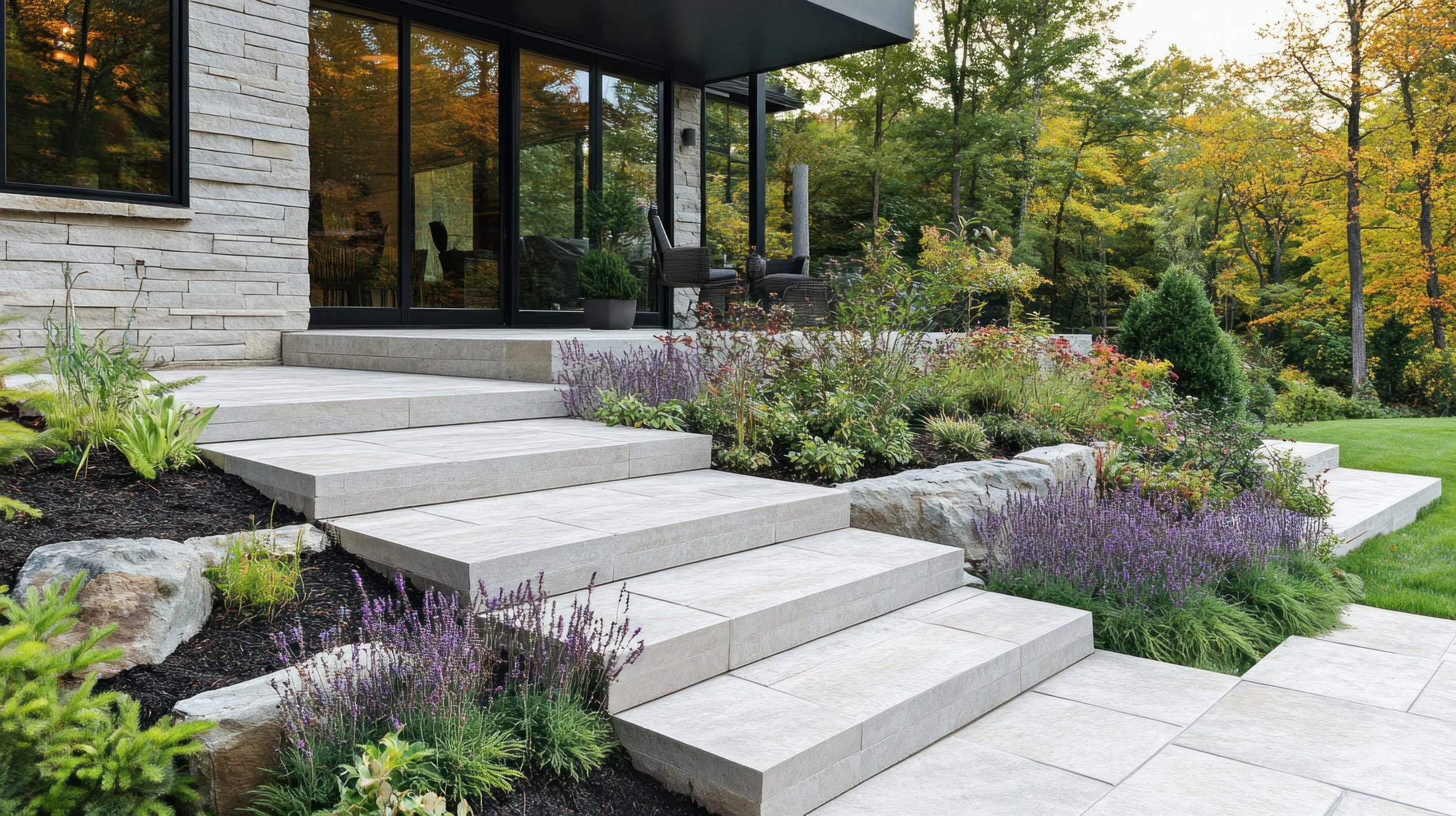 Front porch with stone steps leading to a modern house with glass patio doors, surrounded by landscaped garden with various plants and trees