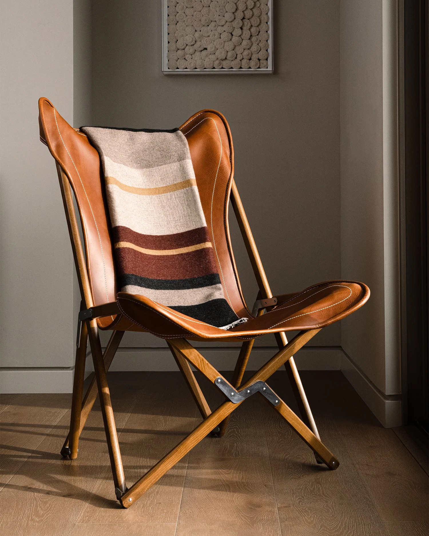 A leather and wood lounge chair with a patterned throw draped over it, placed near a wall and a window with sunlight casting shadows on the wooden floor.