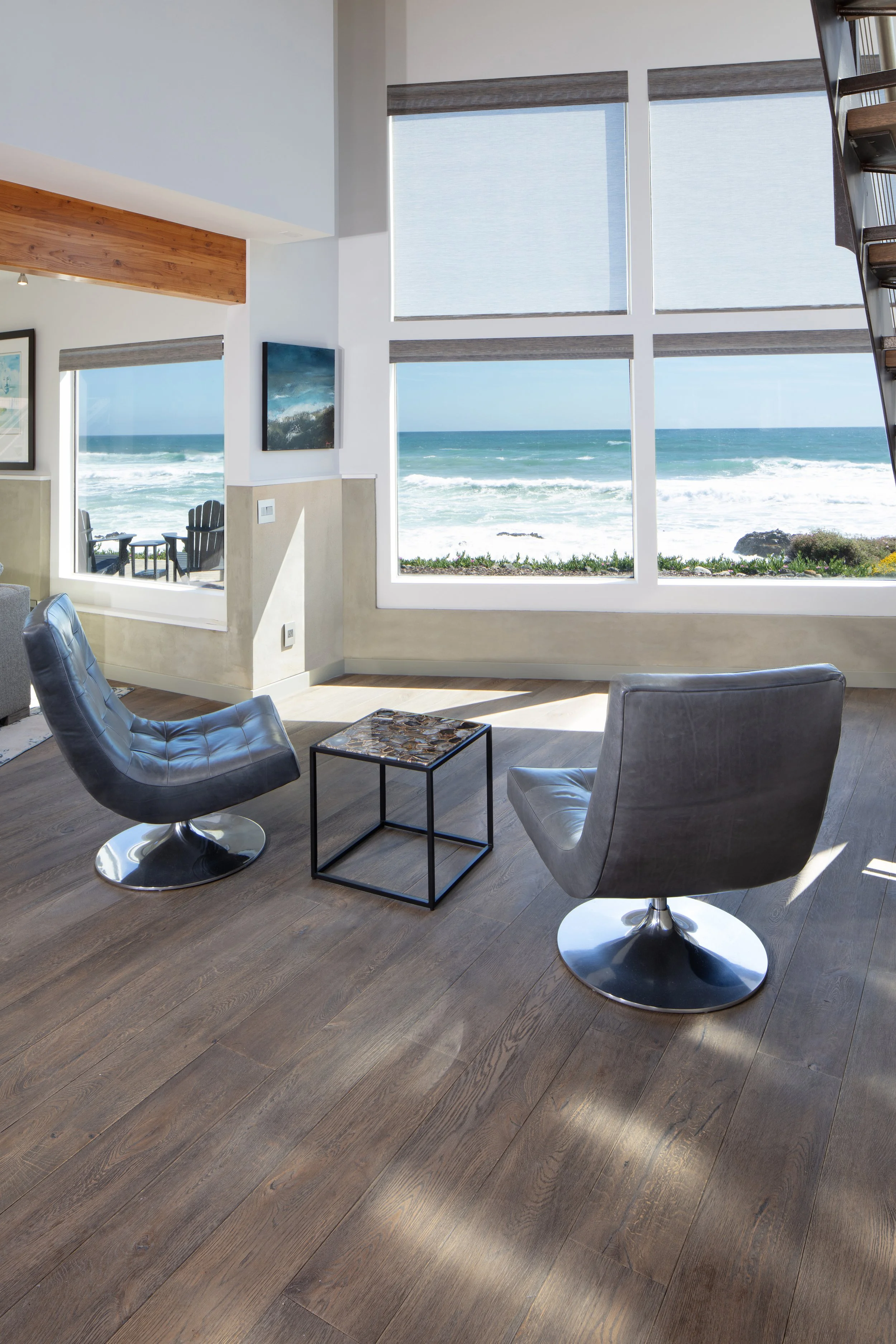 Living room with large windows overlooking the ocean, two black modern chairs, a small table, and ocean scenery outside.