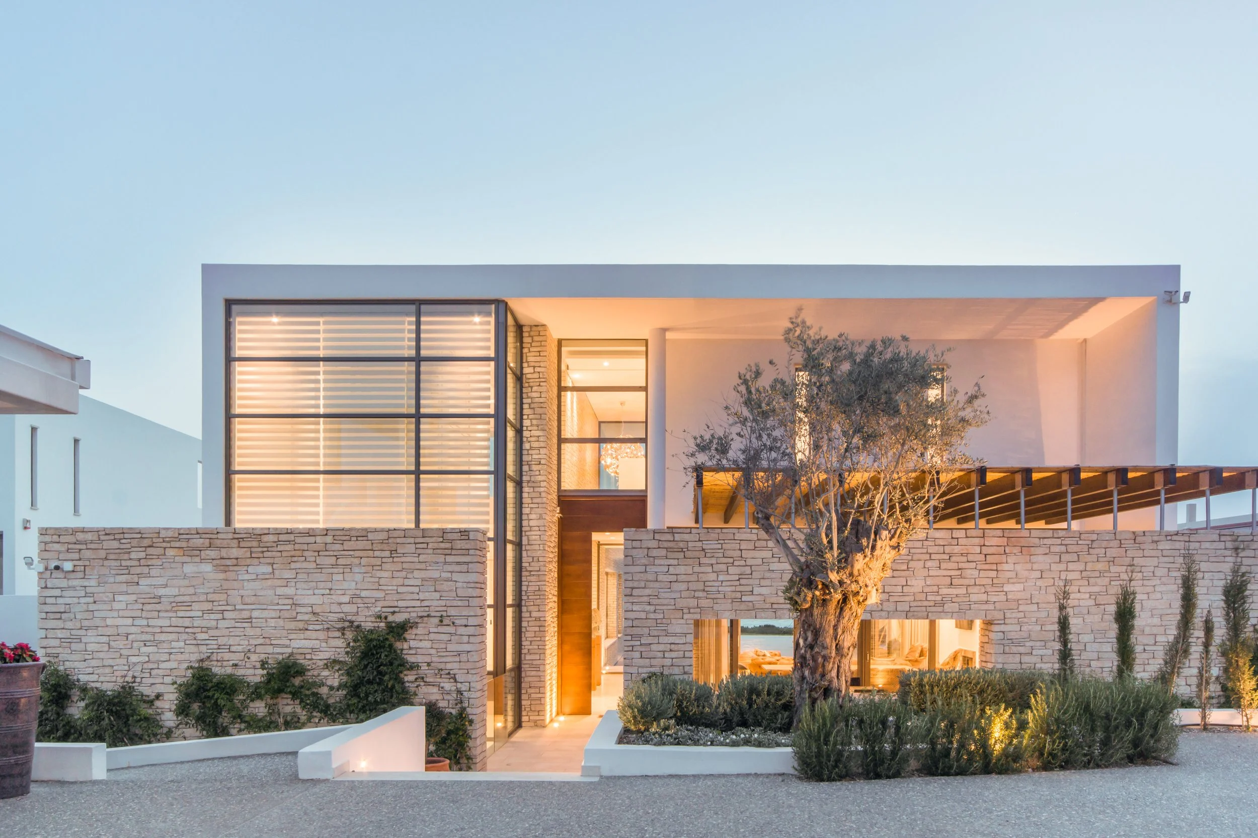 Modern two-story house with dry stack stone and white exterior, large glass windows, and a landscaped front yard with a tree and plants, illuminated in soft lighting.