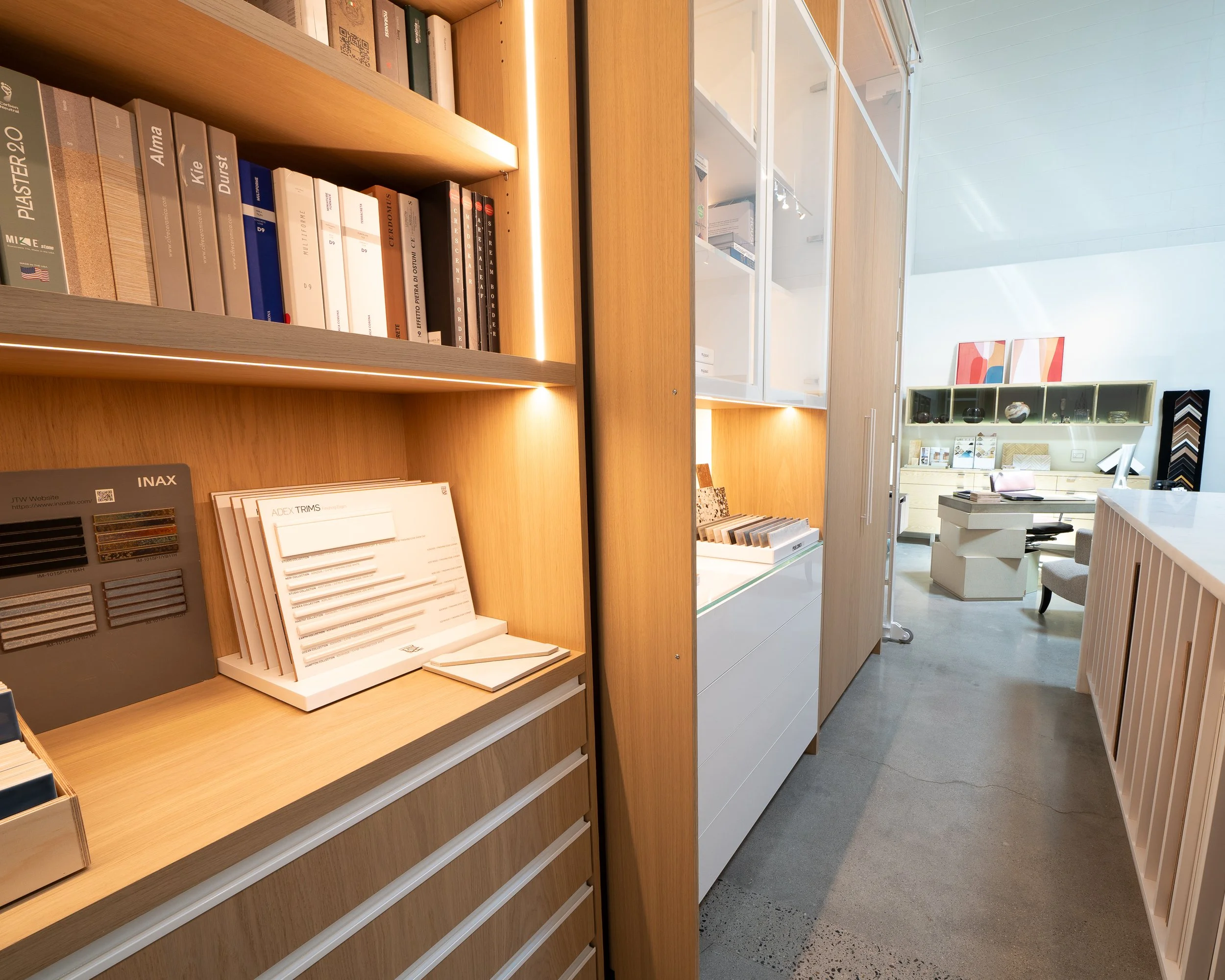 Interior of a modern showroom displaying various interior design samples and furniture, with shelves holding books, fabric swatches, and sample boards.