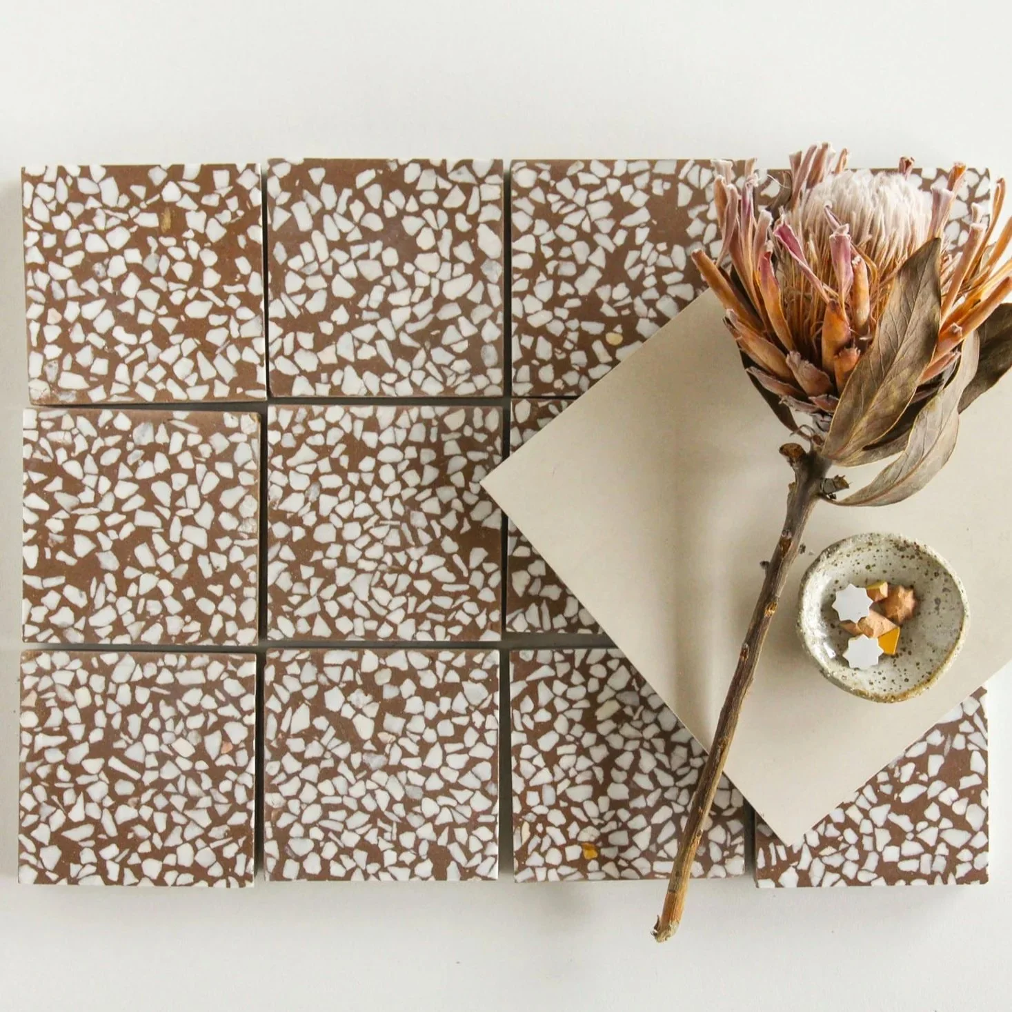 Top view of a white surface with a diagonally placed beige paper, a dried protea flower with pink and brown petals, a speckled ceramic cup containing salt and pepper shakers, and nine terrazzo tiles with brown and white speckled pattern.