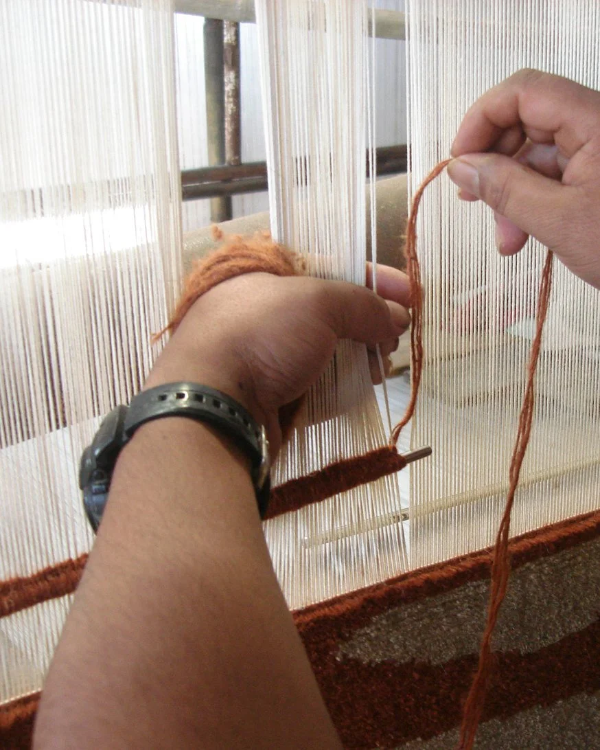 Person hand knotting a carpet with brown yarn on a loom.