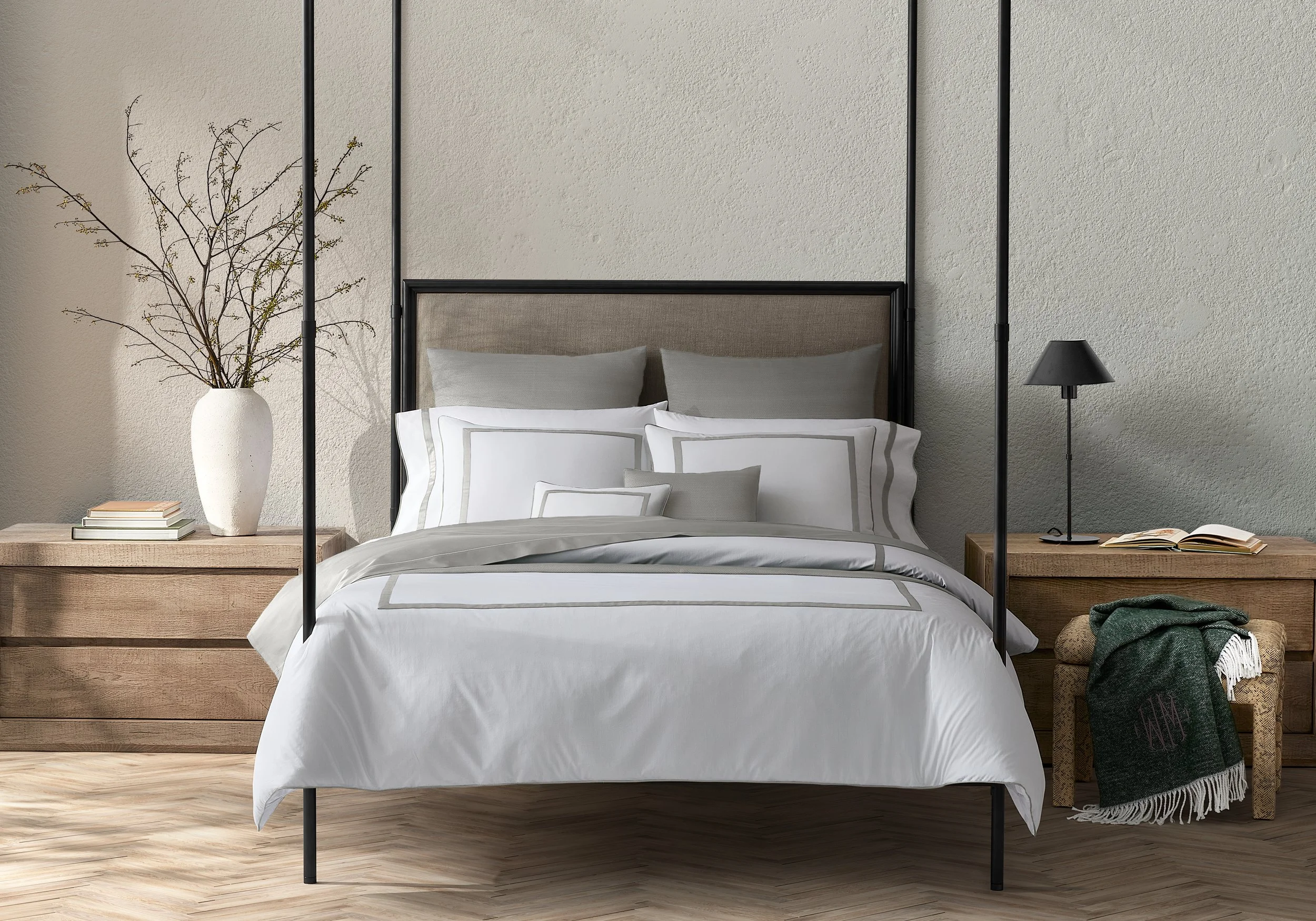 A neatly made bed with white and gray bedding, wooden nightstands on each side, one with a white vase holding branches, the other with a black lamp and an open book, a throw blanket on a small wooden stool, all against a textured gray wall.