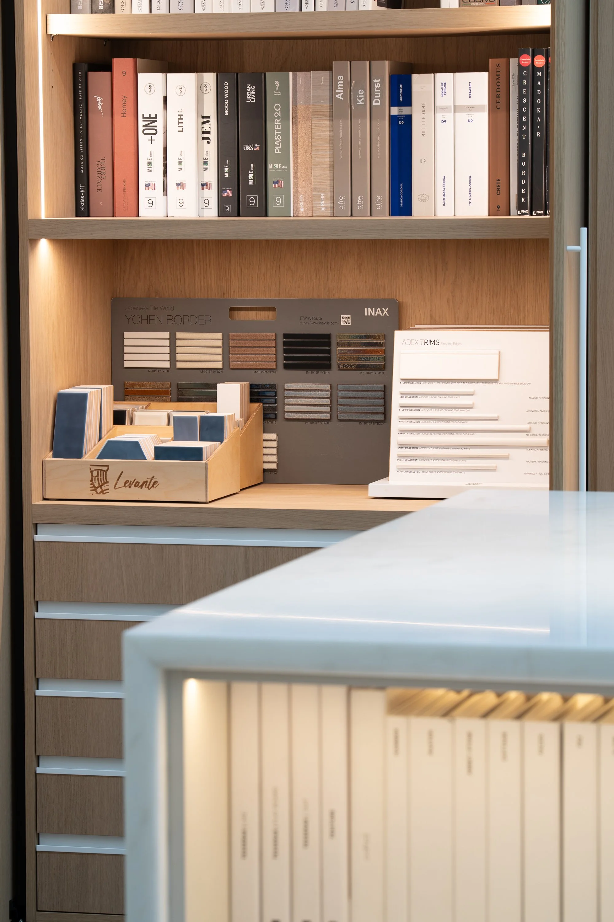 Interior of a wooden shelving unit displaying architecture and design books on the top shelf, sample tiles and trim samples on the middle shelf, and a stack of white books or catalogs on the bottom right.