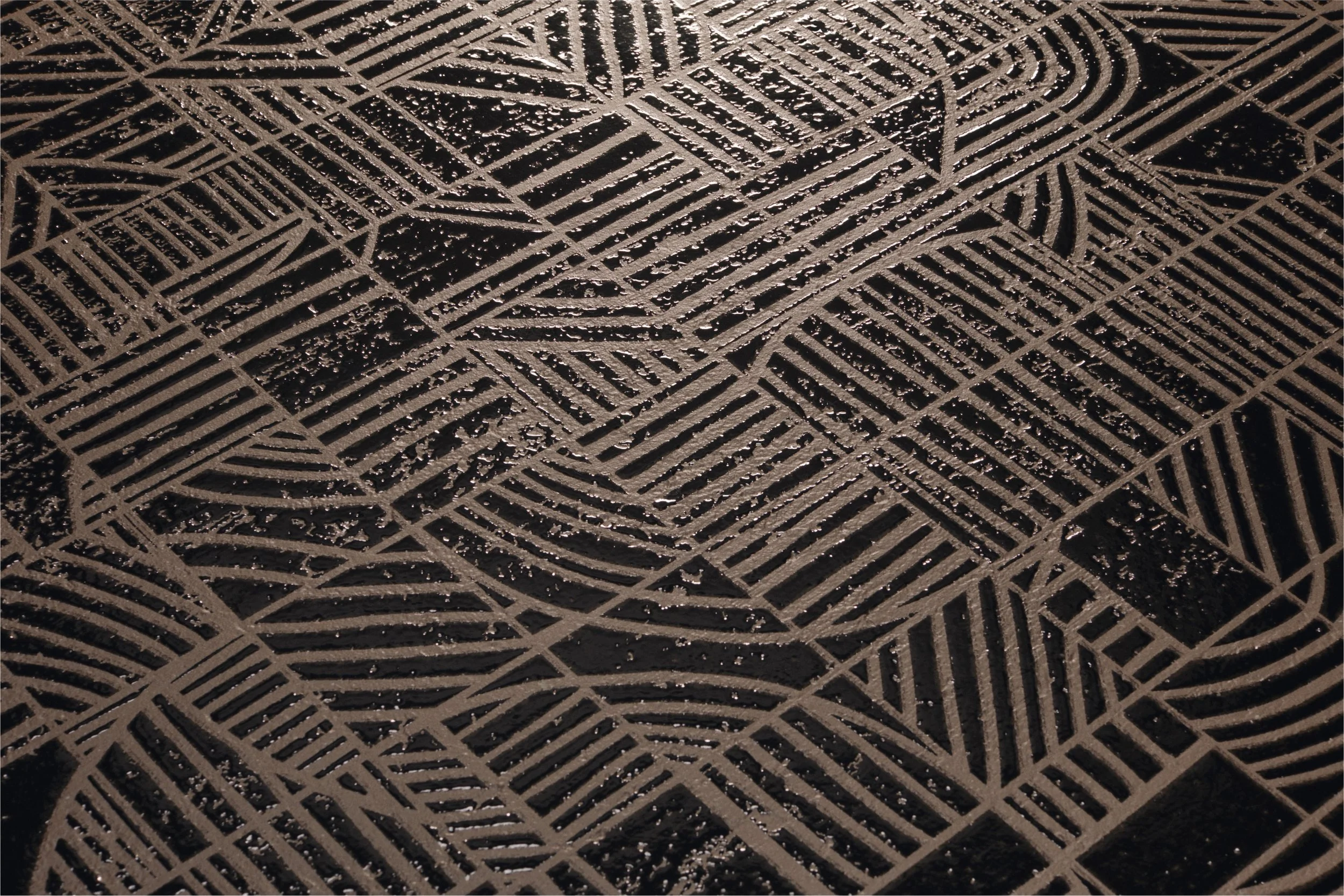 Close-up of a black textured surface with intricate geometric and linear patterns in brown or tan. The patterns include lines and shapes forming squares, rectangles, and curved structures.