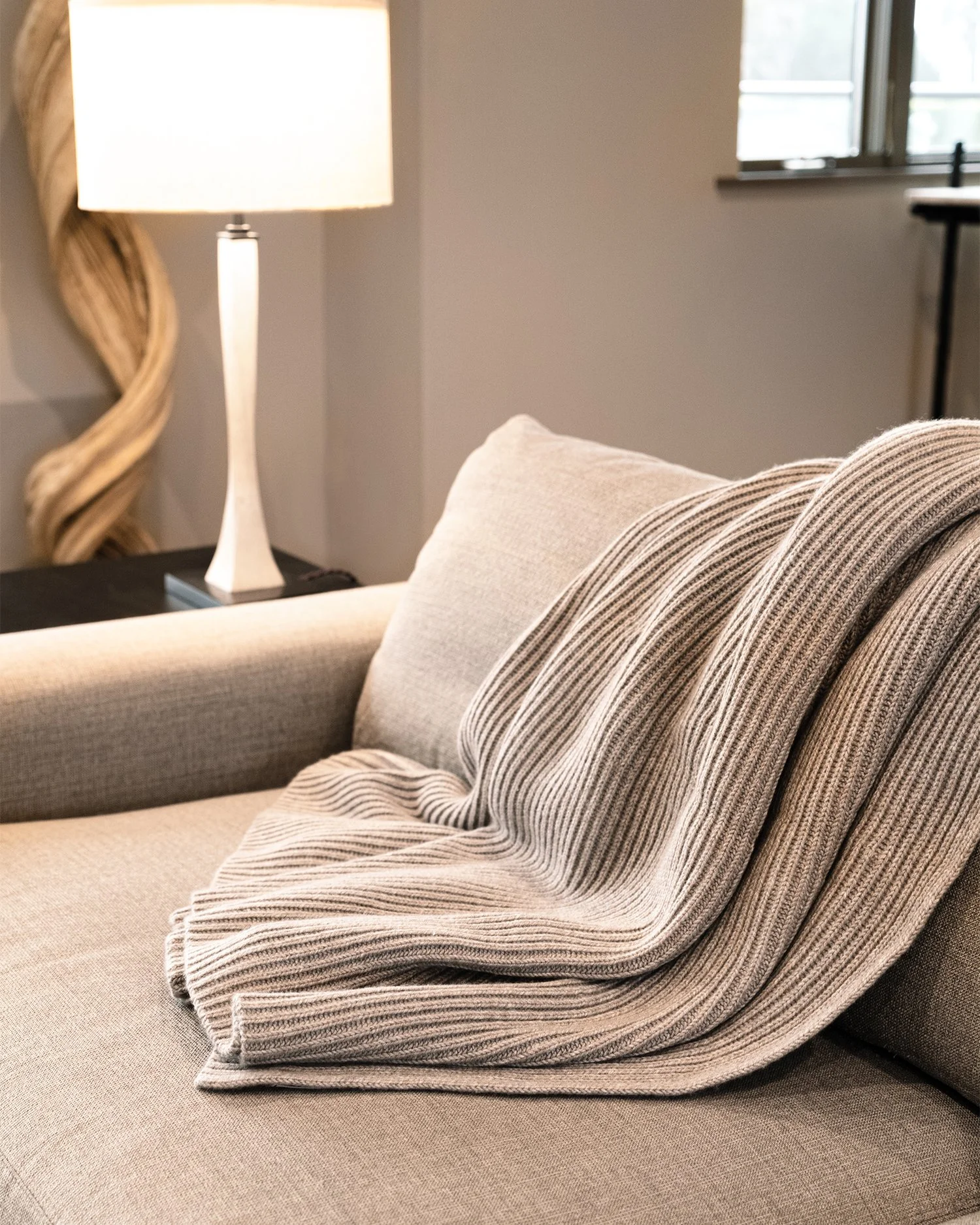 Close-up of a beige fabric sofa with a matching striped blanket and a cushion, with a lamp and window in the background.