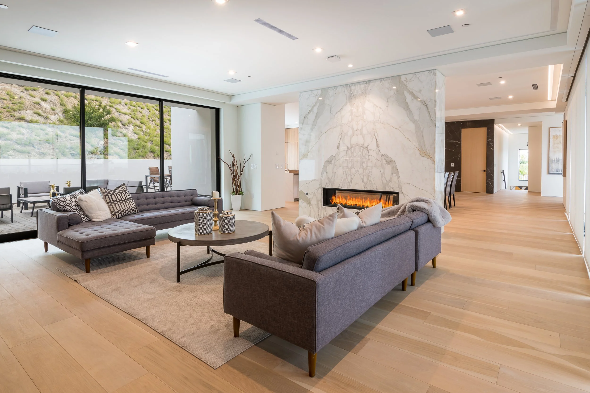 Modern living room with gray couches, a fireplace, wooden floors, and large sliding glass doors leading to an outdoor patio.