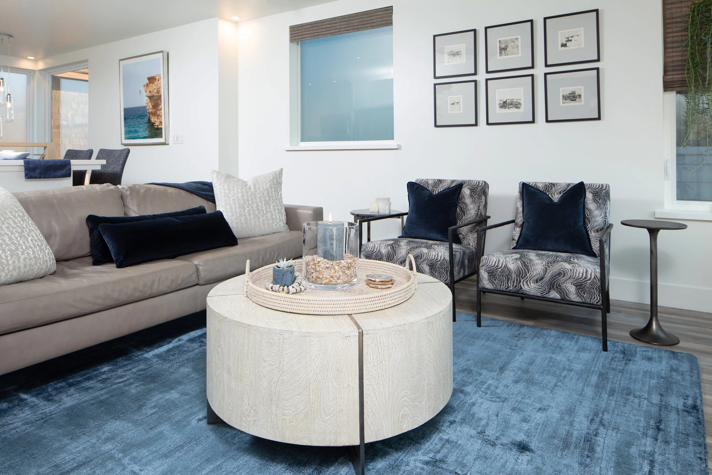 Living room with beige sofa, patterned armchairs with black pillows, a round light-colored coffee table with candles and decorative items, a blue area rug, and framed artwork on the white walls.