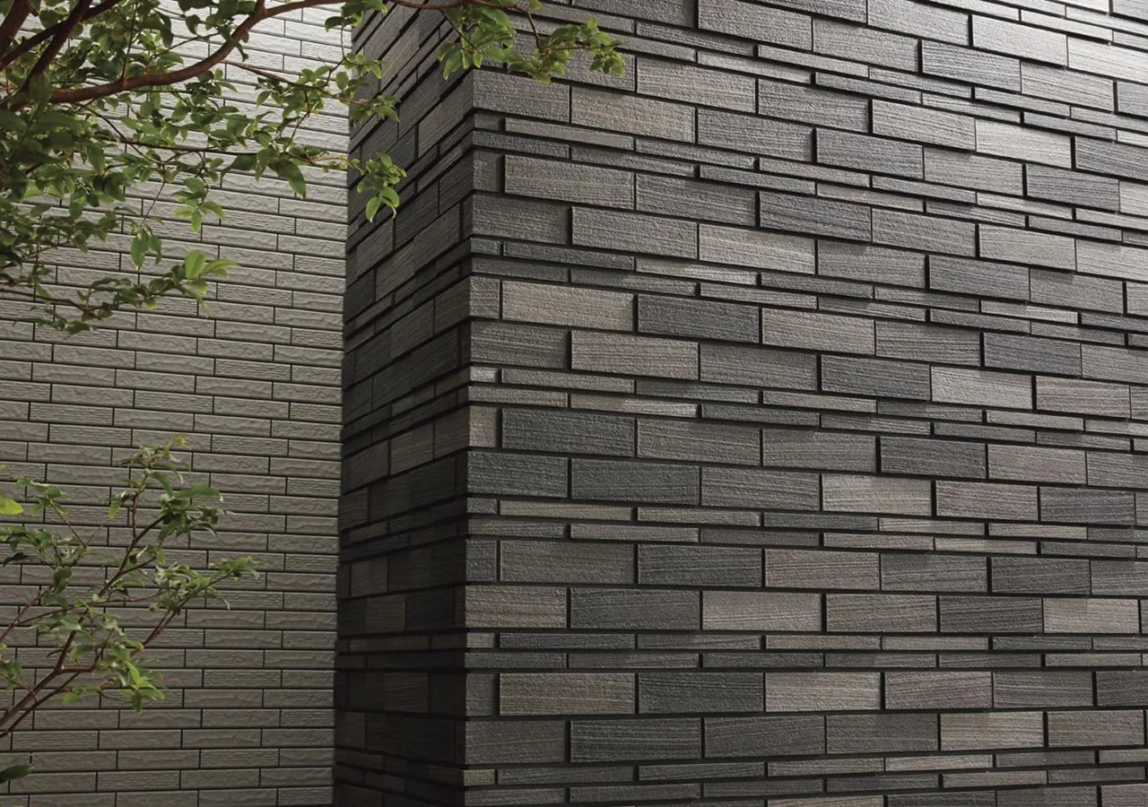 Close-up of a brick wall with dark gray bricks and a corner section, with some green leaves and branches in the foreground.