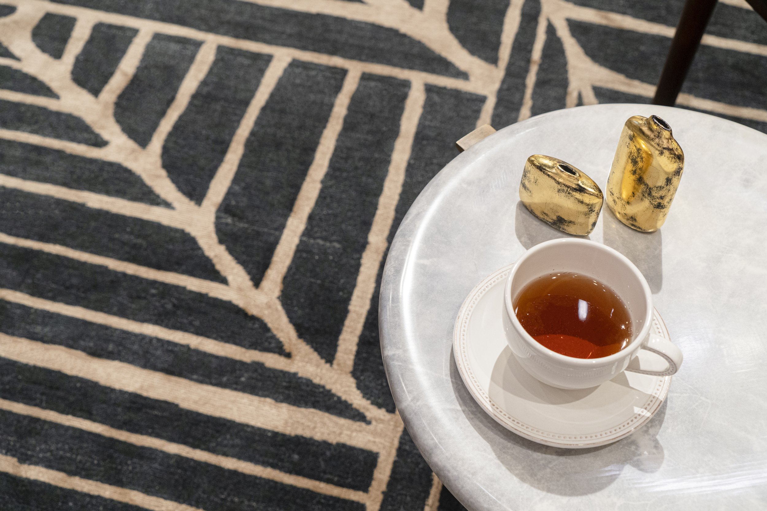 A white cup of tea on a saucer, with two gold-colored decorative objects on a marble table, next to a patterned carpet.