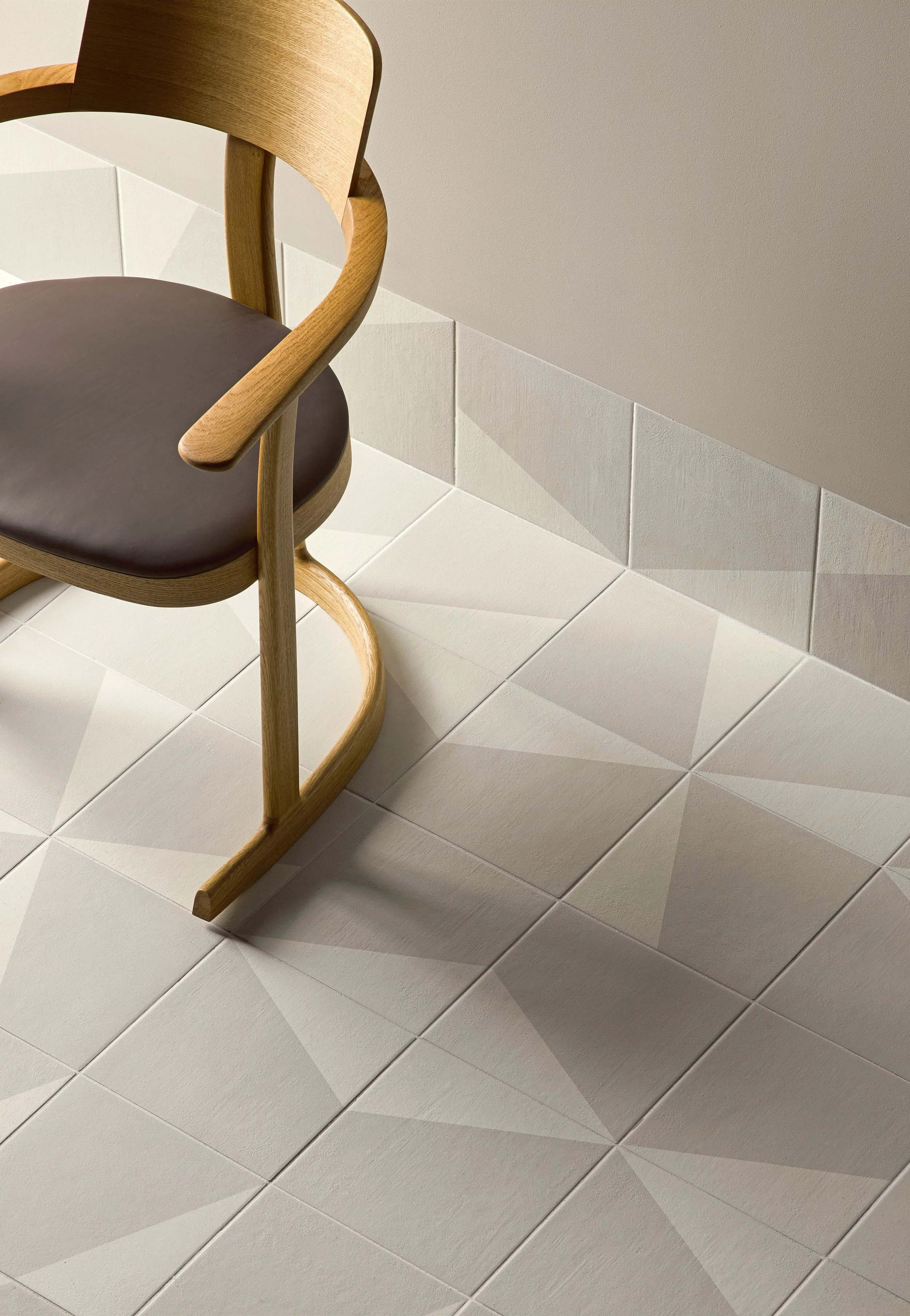 A wooden chair with a dark brown cushioned seat sits on a tiled floor with geometric patterns, near a plain beige wall.