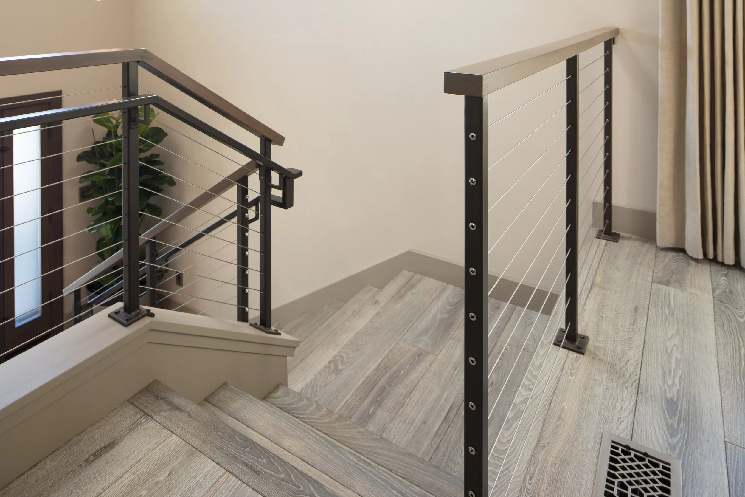 Interior view of a staircase with modern black metal and wood handrails, beige walls, light wood flooring, a potted plant by a window, and a curtain on the right side.