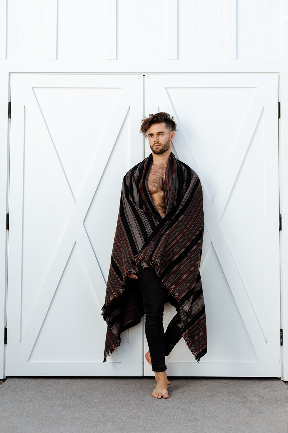 A man with styled hair and facial hair standing barefoot in front of white double doors, wrapped in a striped shawl or blanket that covers his shoulders and flows down to his legs.